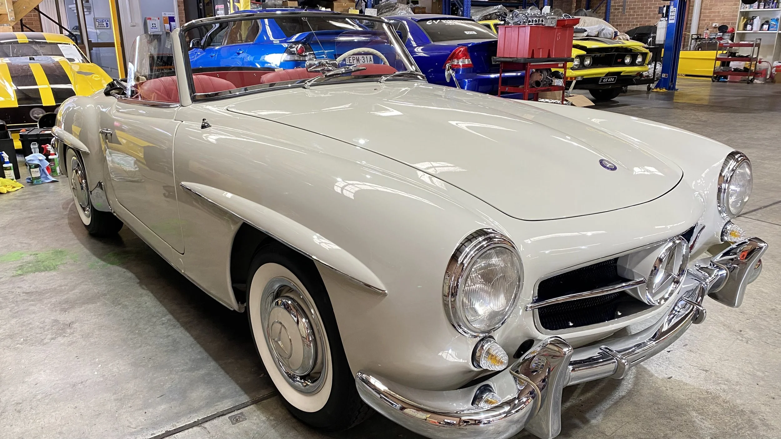 A vintage white convertible car with a red interior parked inside a garage or workshop, with multiple other cars and automotive tools visible in the background.