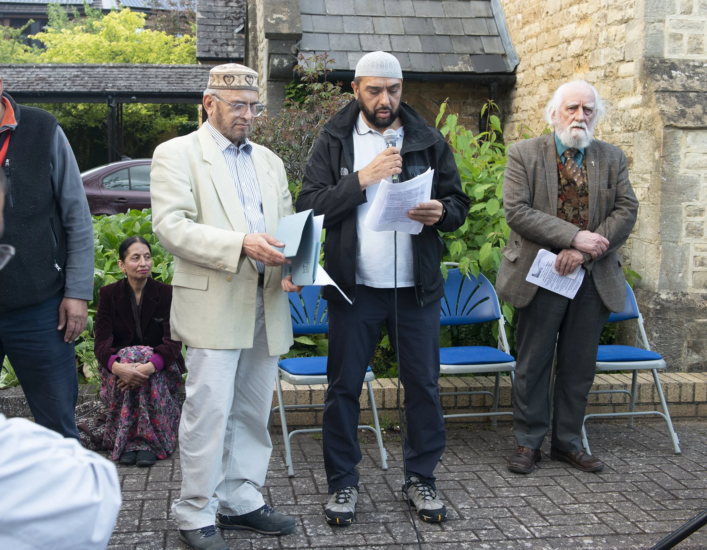 A group of people attending an outdoor event, some standing and some sitting. The man in the center is speaking into a microphone, holding papers, and wearing casual outdoor clothing including a black jacket and a gray cap. The man to his right has a