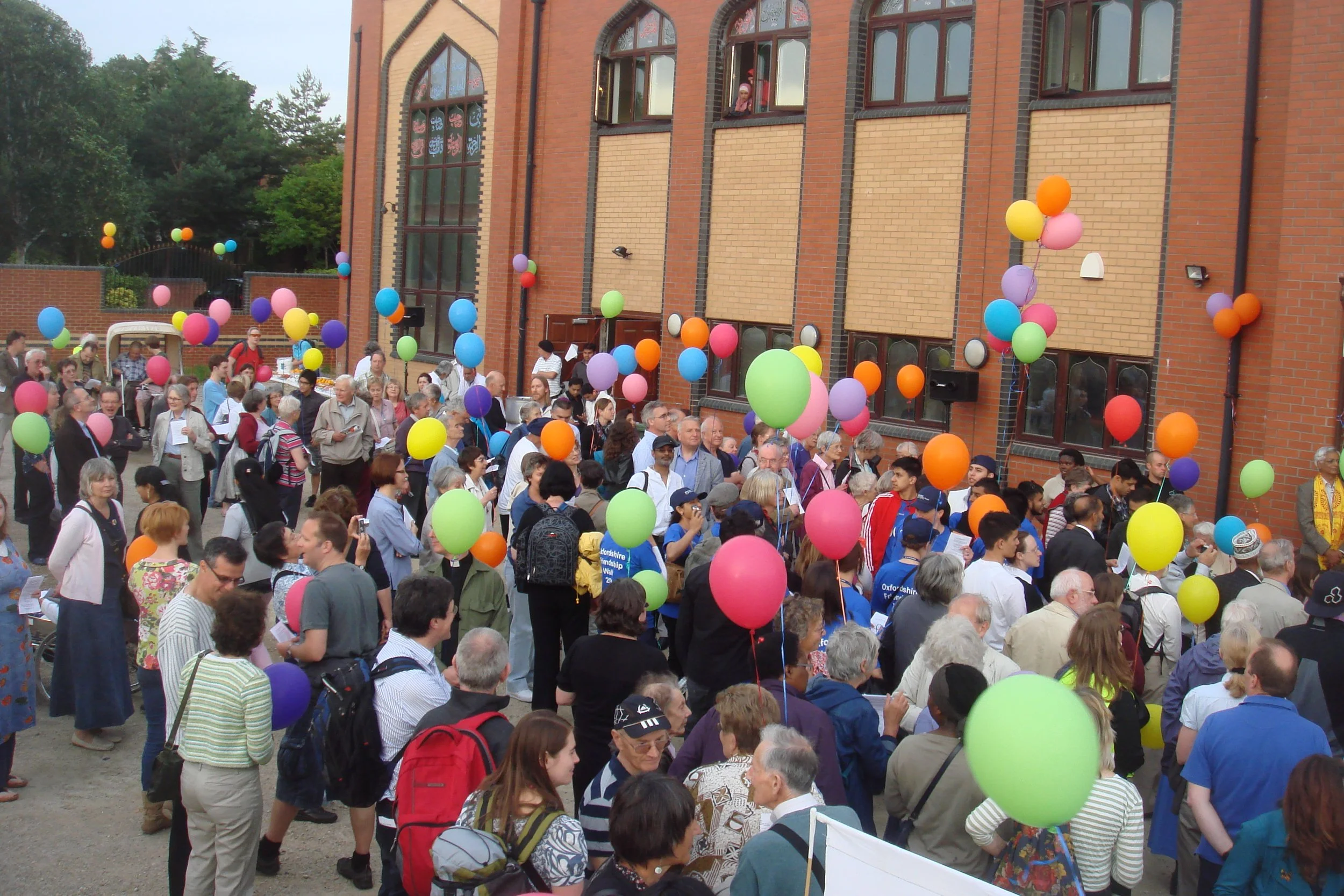A crowd of people gathered outside a brick building with large windows, decorated with colorful balloons.