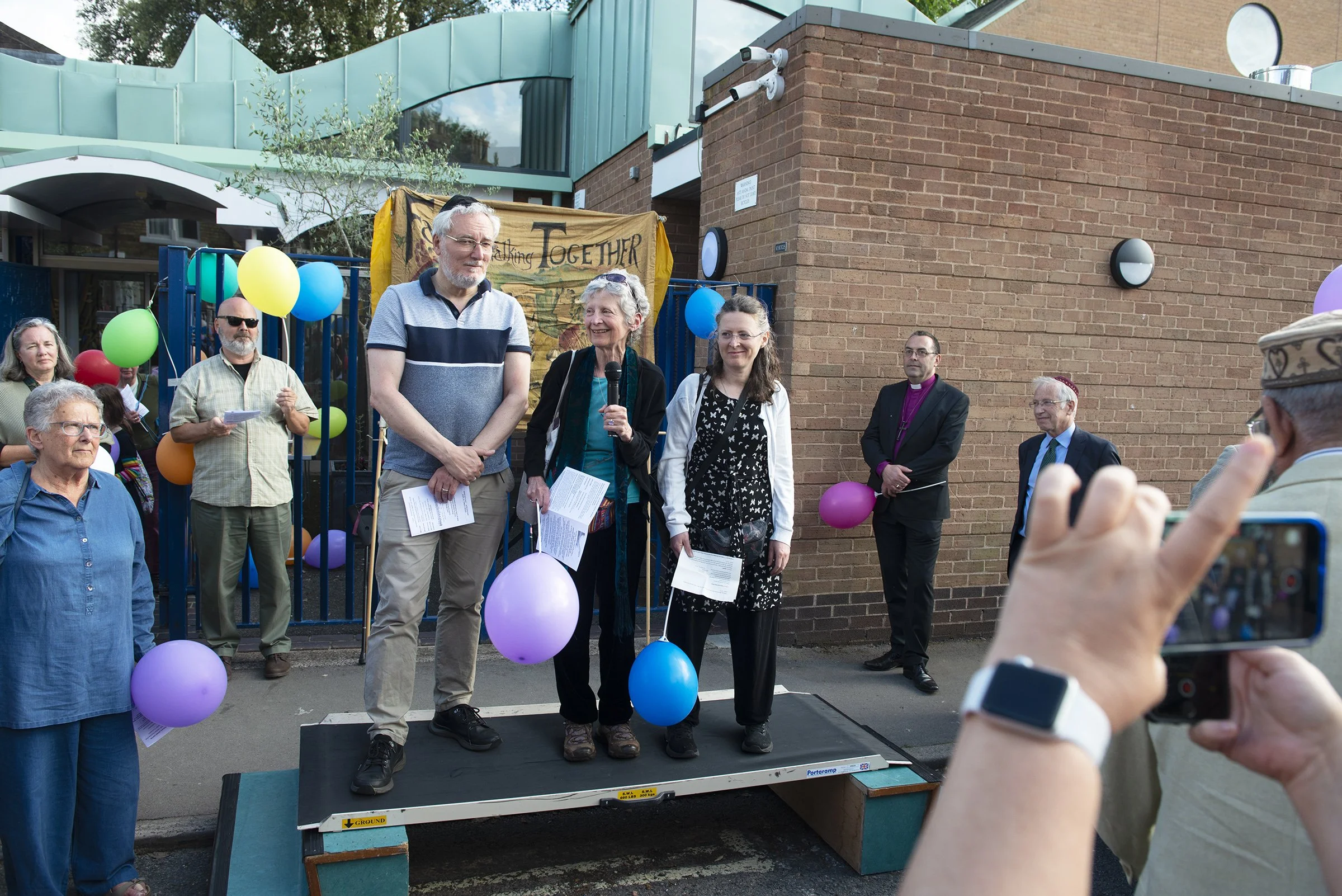 A group of people gathered outdoors during a community event, with some standing on a small platform. The scene includes colorful balloons, a man speaking into a microphone, and a background banner with the word 'Together.' The event appears festive,