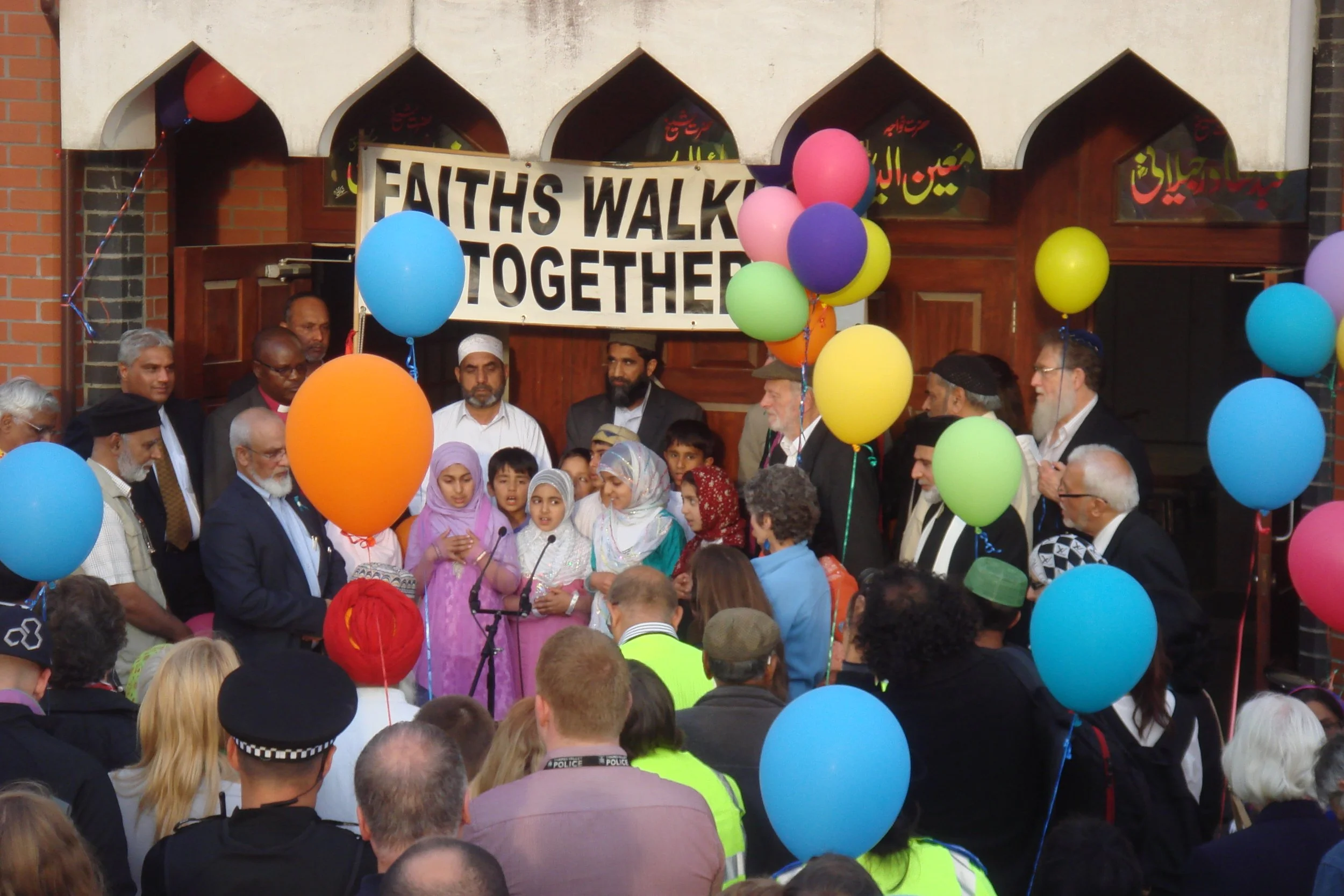 A diverse crowd gathered outside a building decorated with colorful balloons and a large banner reading 'FAITHS WALK TOGETHER.' People, including children and adults, participate in a multicultural event, some wearing traditional clothing, with the s