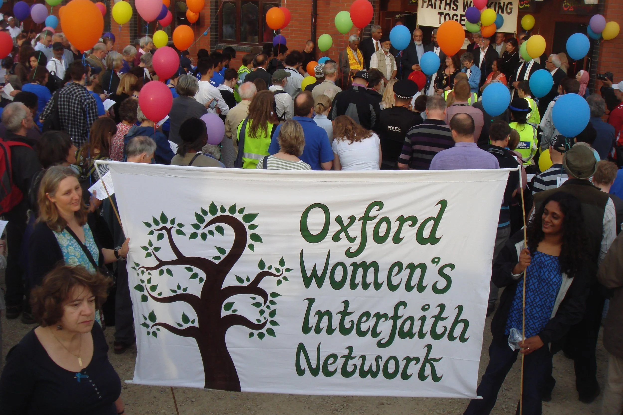 A large crowd of people gathered at a rally, holding a banner that reads 'Oxford Women's Institute.' Many people are holding colorful balloons, and there are several individuals wearing brightly colored vests. The event appears lively and celebratory