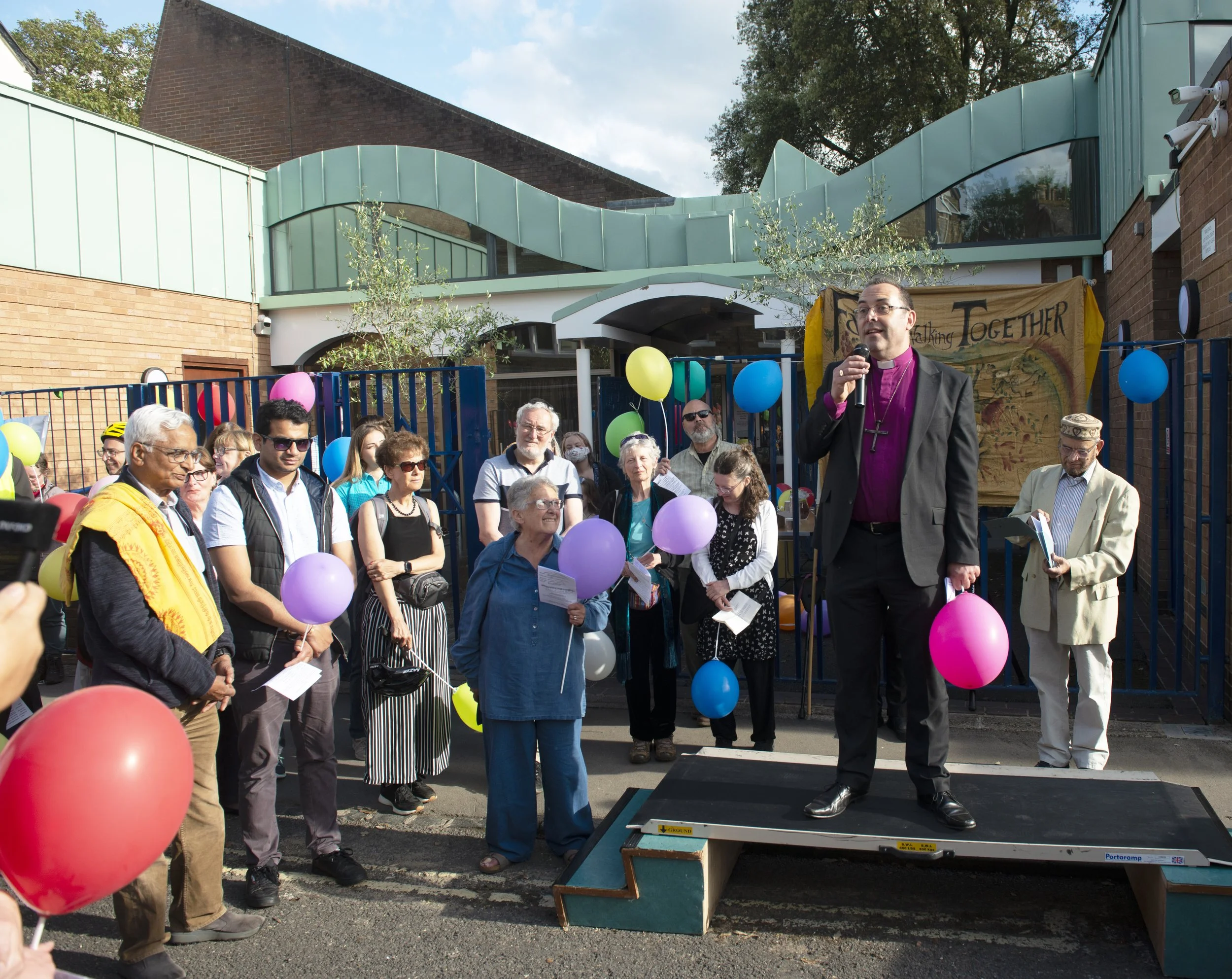 A crowd of people gathered outdoors for a community event, with a man in clerical attire speaking into a microphone while standing on a small platform. The event is decorated with colorful balloons.