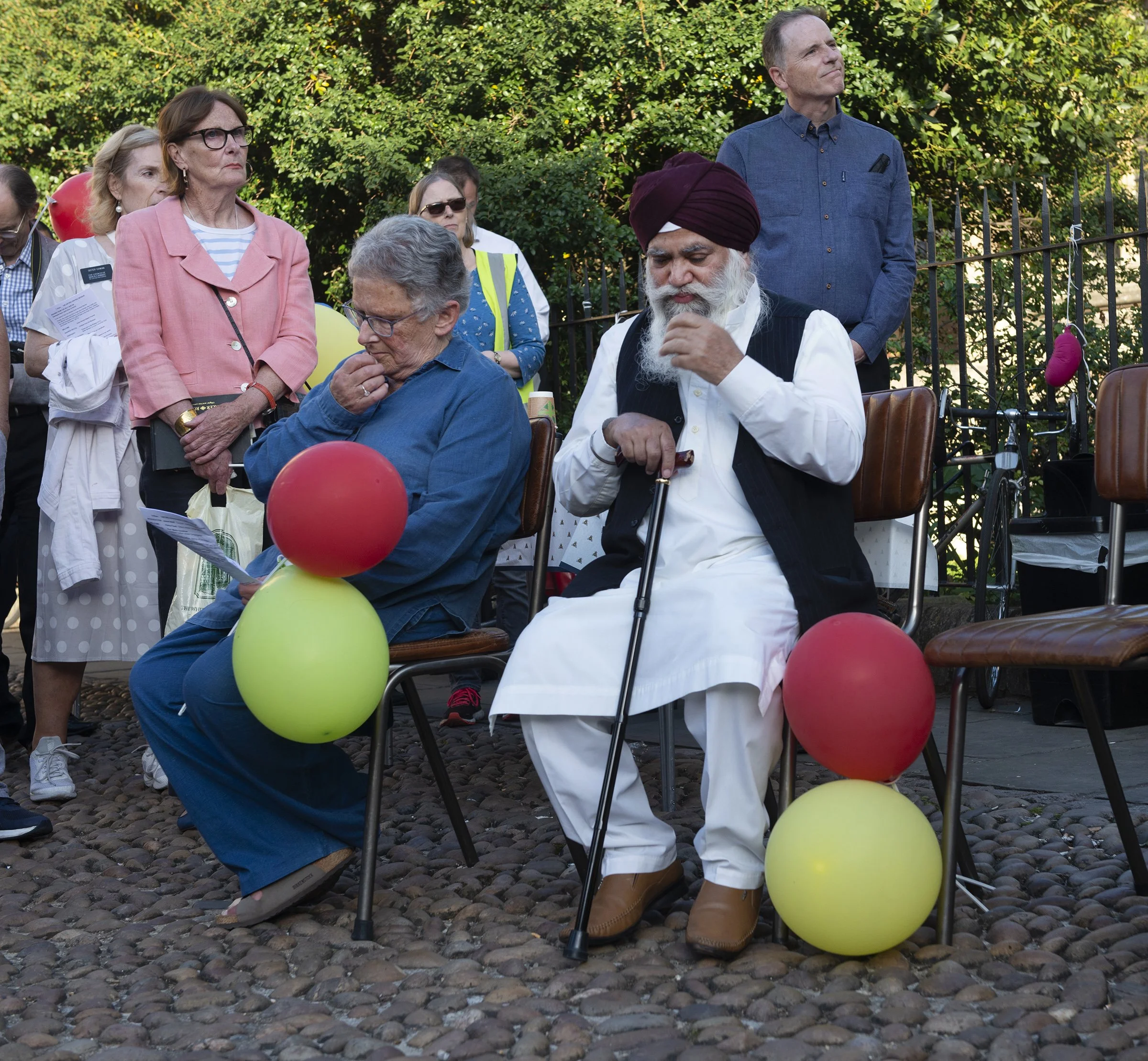Group of five elderly people gathered outdoors, some standing and some sitting, with colorful balloons intertwined among their chairs. The background features lush green foliage. One man with a long white beard and wearing a purple turban appears to 