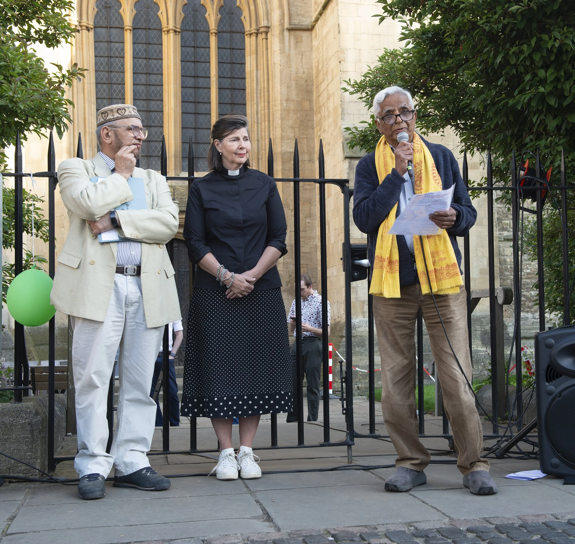 Three people standing outside in front of a building with large windows, next to a black metal fence and green bushes. The person on the right is speaking into a microphone while holding papers, the person in the middle is a woman in a black outfit, 