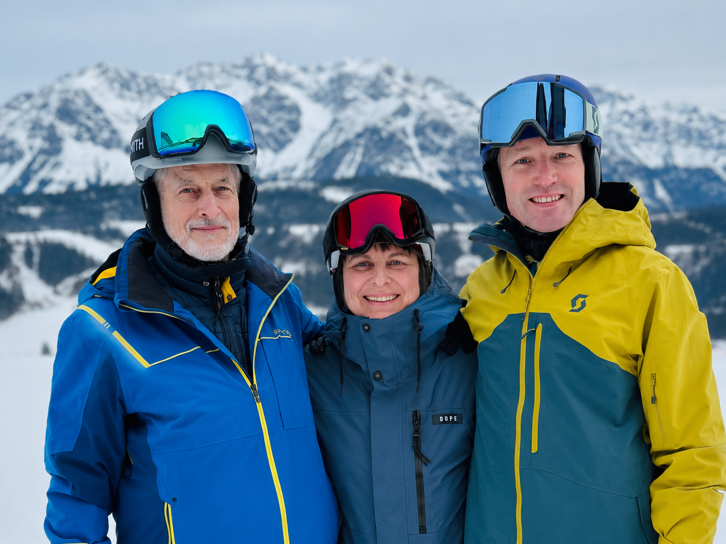 Drei Menschen in Skiausrüstung lächelnd vor schneebedeckten Bergen.