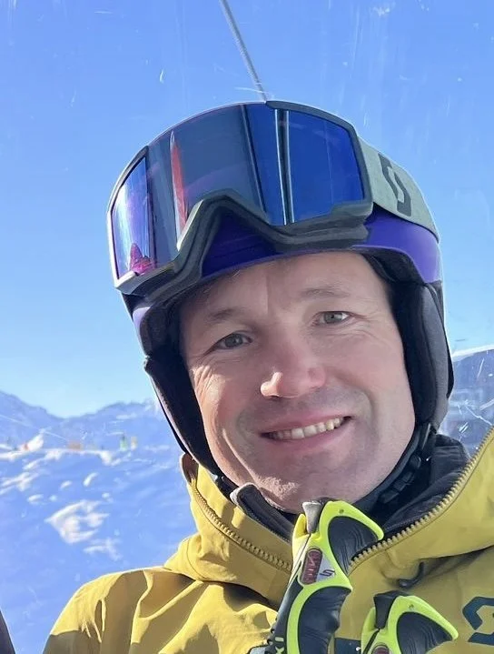 Mann trägt einen Skihelm und eine Skibrille, trägt eine gelbe Skijacke und befindet sich auf einer Skipiste mit verschneiten Bergen im Hintergrund.