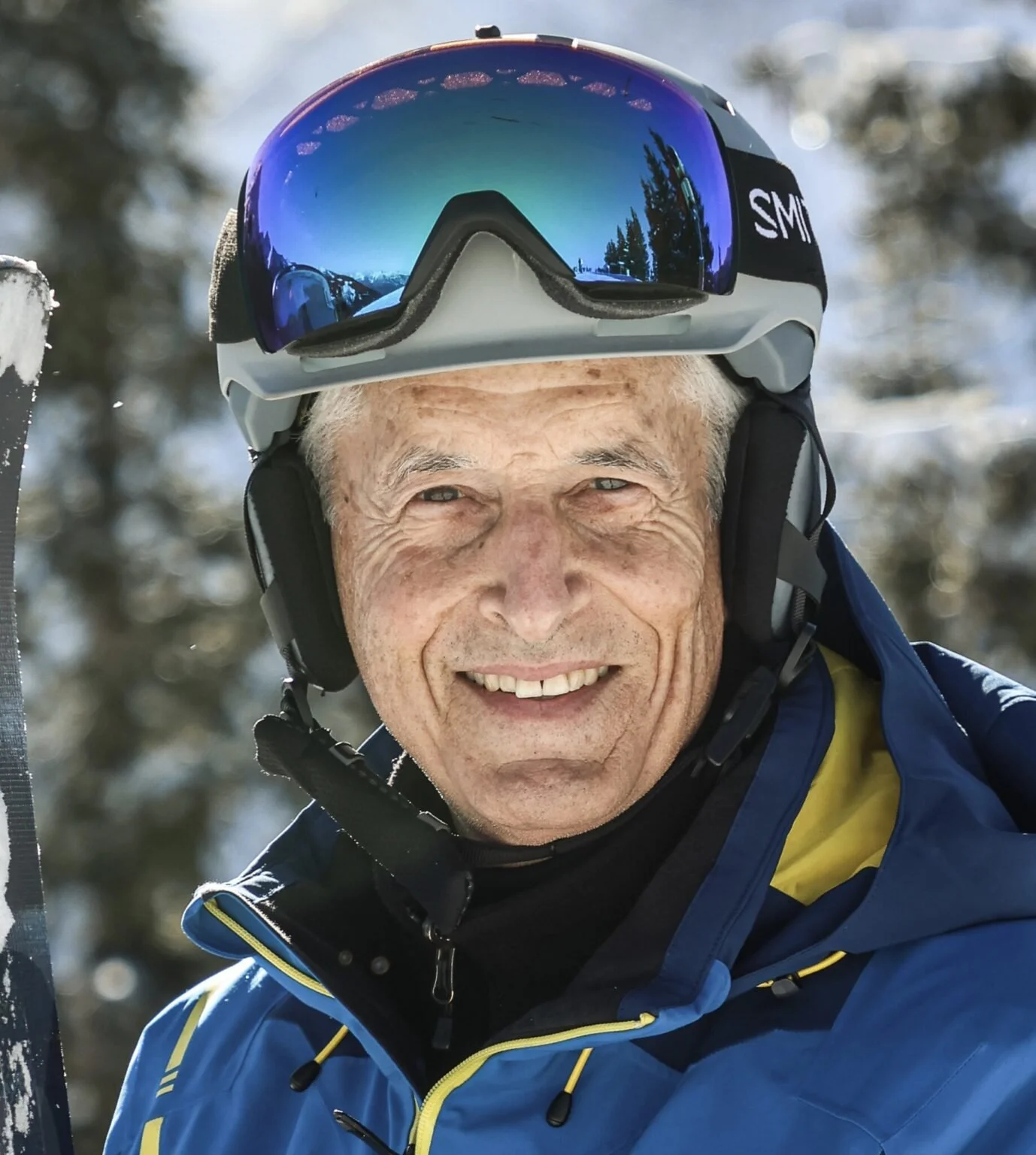 Ein älterer Mann trägt einen blauen Skianzug, eine Skibrille auf dem Kopf und einen Helm, und lächelt in die Kamera. Im Hintergrund sind verschneite Bäume, es sieht nach einem Wintersport in den Bergen aus.