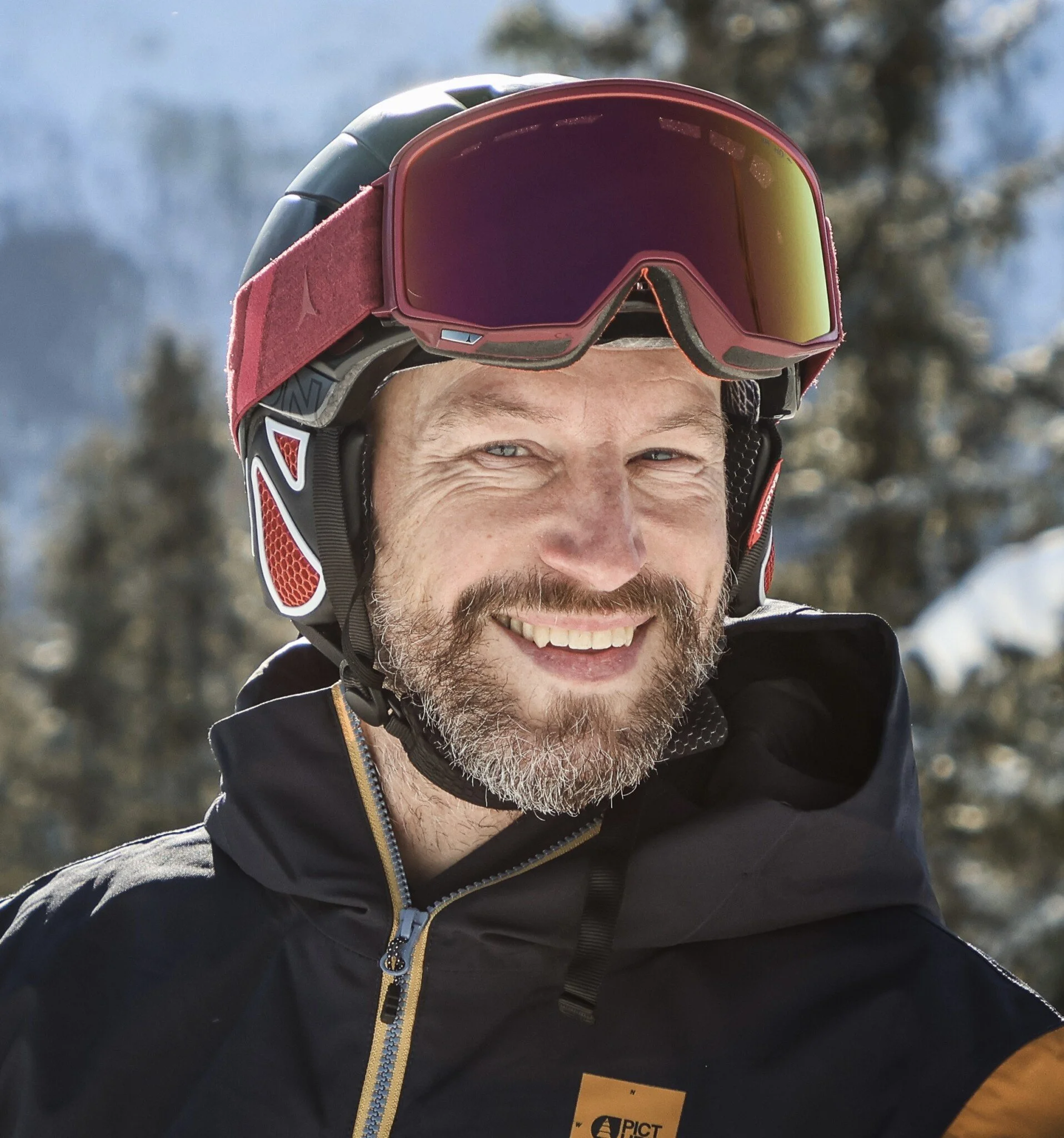 Porträt eines lachenden Mannes mit Glatze und Bart, trägt Skibrille, rote Mütze und schwarze Jacke, draußen mit Bäumen im Hintergrund.
