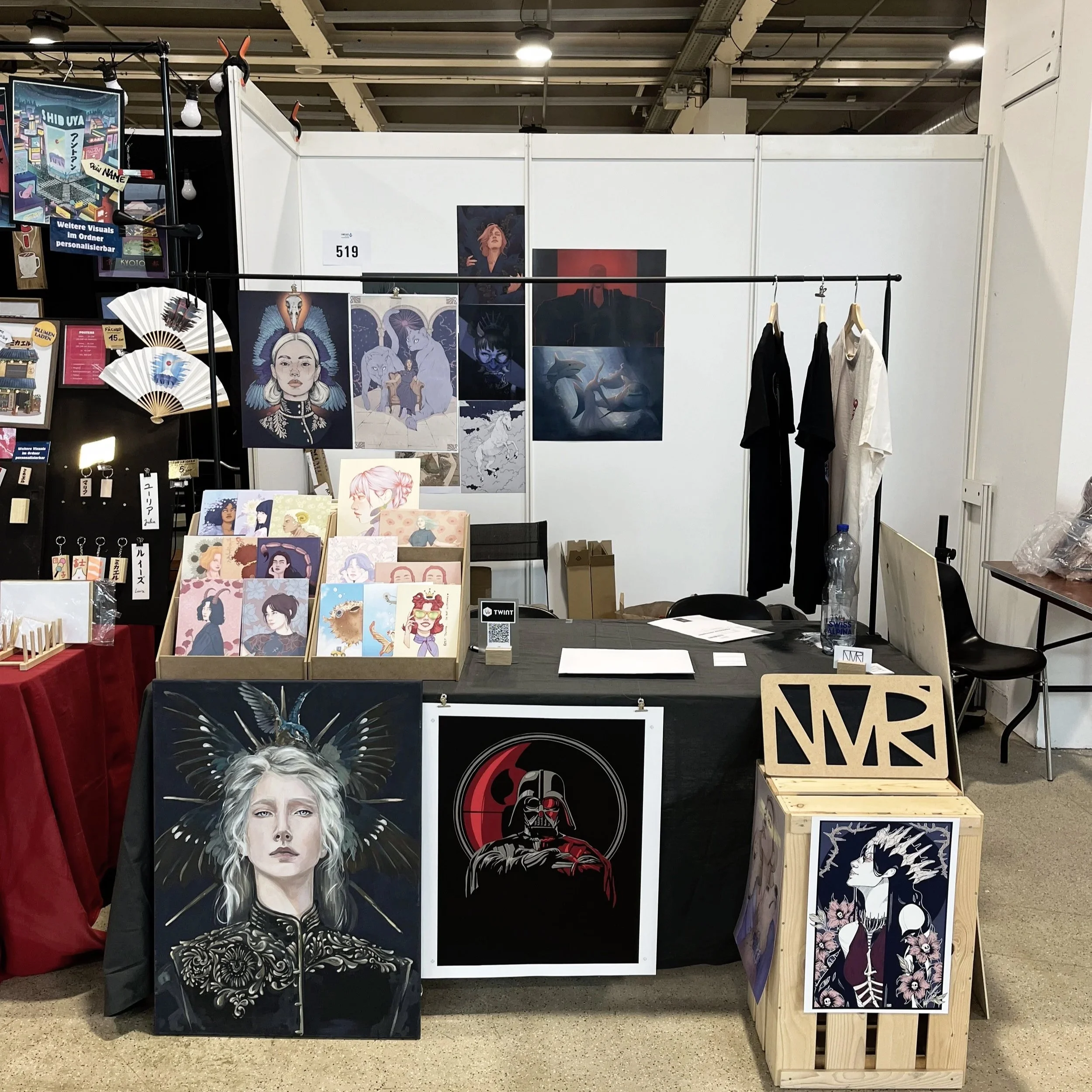 Stände mit Kunstwerken und Postern bei einer Kunstmesse, verschiedene bunte Zeichnungen und Designs von Menschen und Tieren, teilweise mit fantastischen Elementen, hängen an den Wänden und liegen auf Tischen