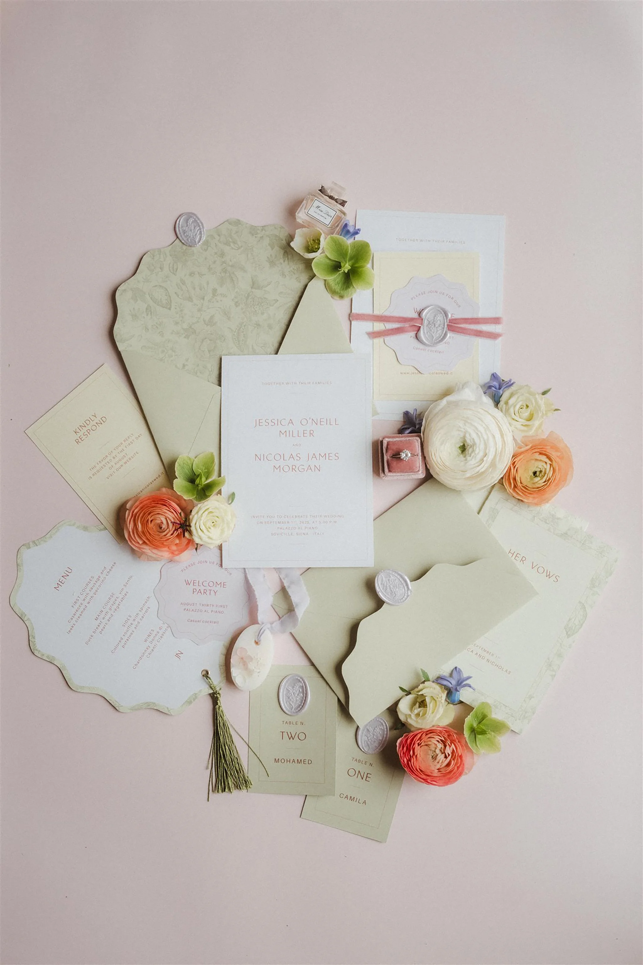 Invito di matrimonio decorato con fiori color pastello, francobolli di ceralacca, biglietti e dettagli eleganti appoggiati su uno sfondo rosa chiaro.