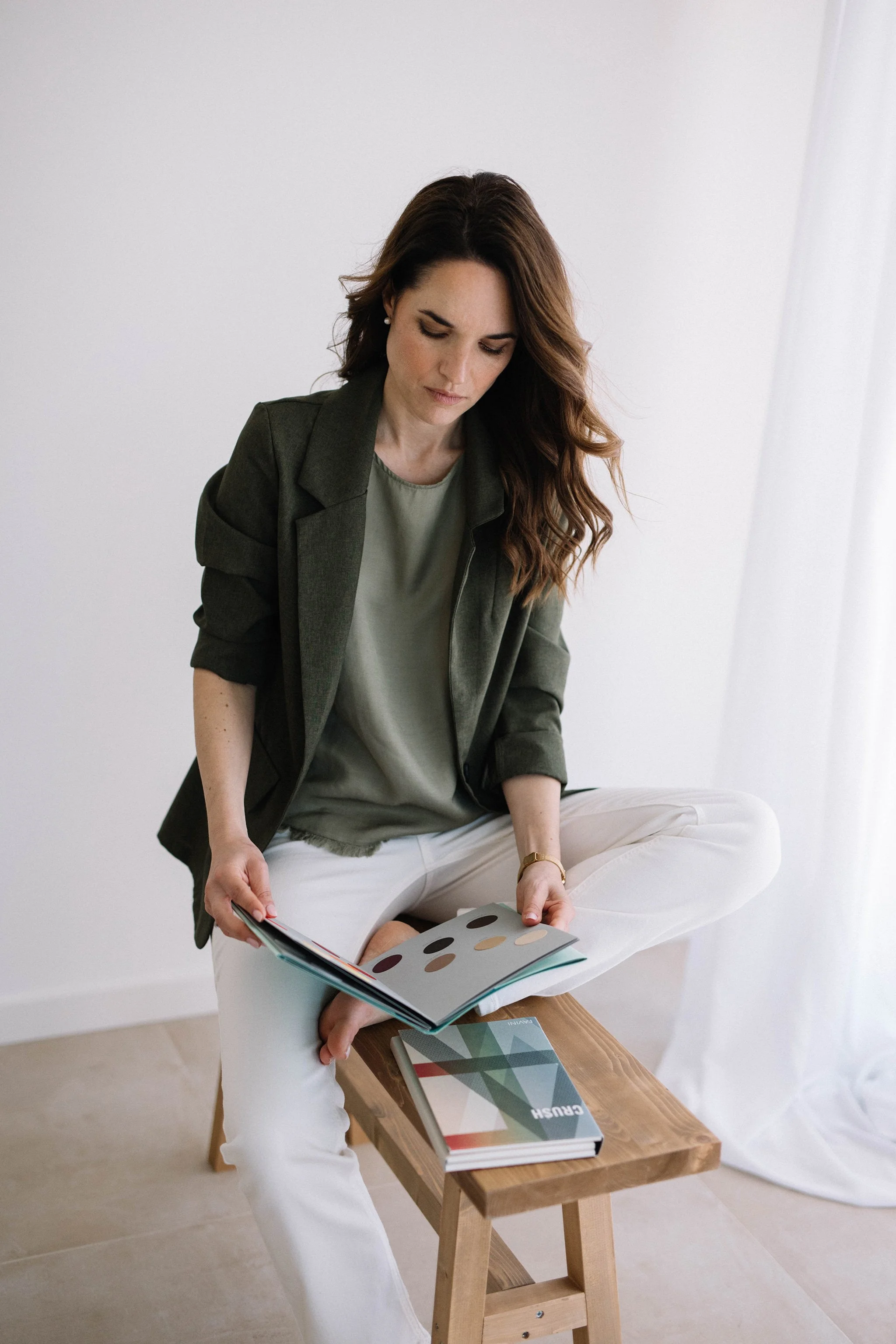 Donna seduta su una sedia di legno, tiene in mano un catalogo di design, indossa una maglietta verde, un blazer grigio e pantaloni bianchi, con una serie di libri e riviste aperte sul tavolo accanto.