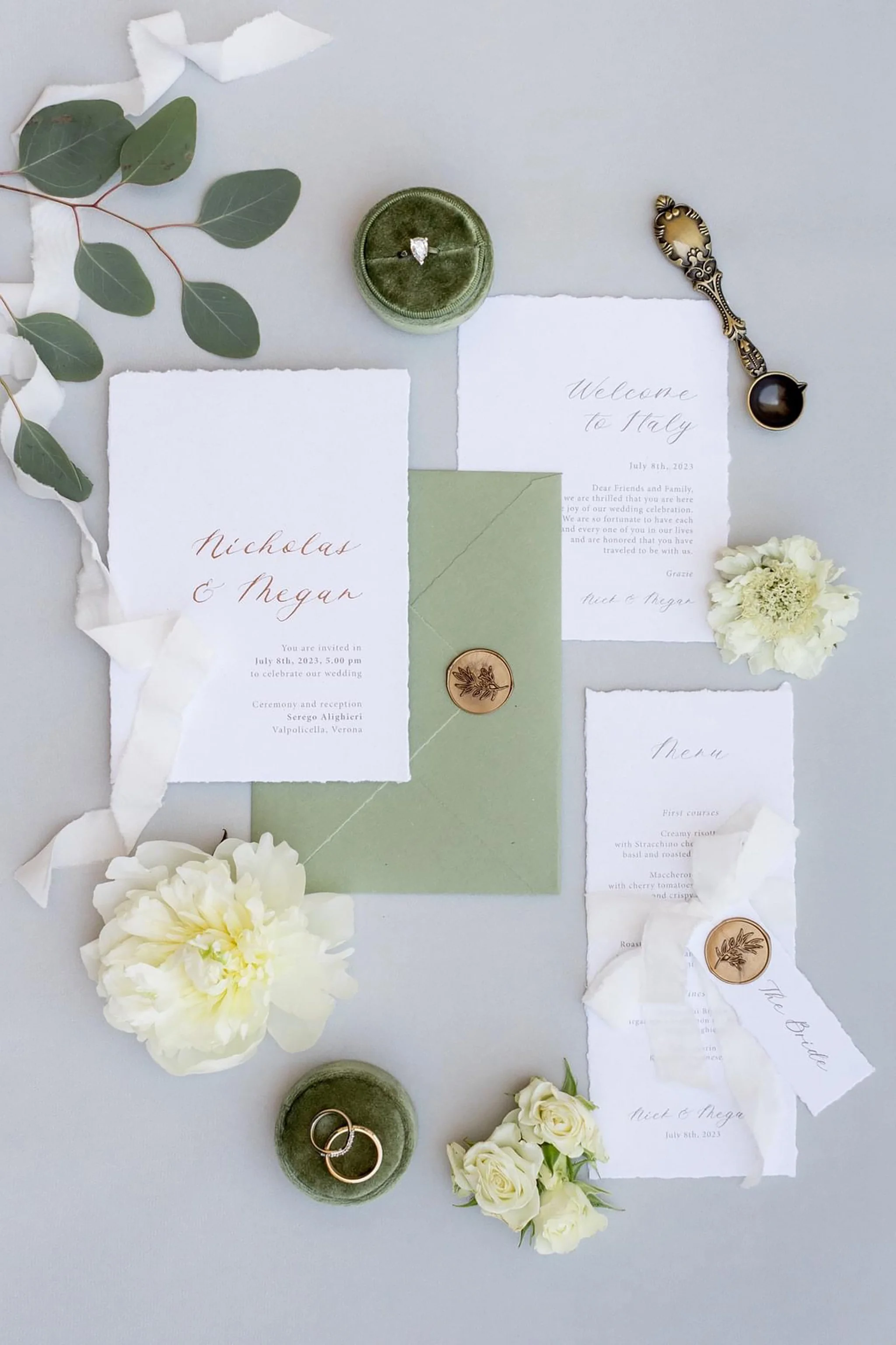 Invito di nozze con fogli di carta bianca, fiori bianchi, foglie verdi, anelli nuziali, una scatola di velluto verde con un anello di diamanti, una chiave antica decorativa e biglietti con messaggi in italiano