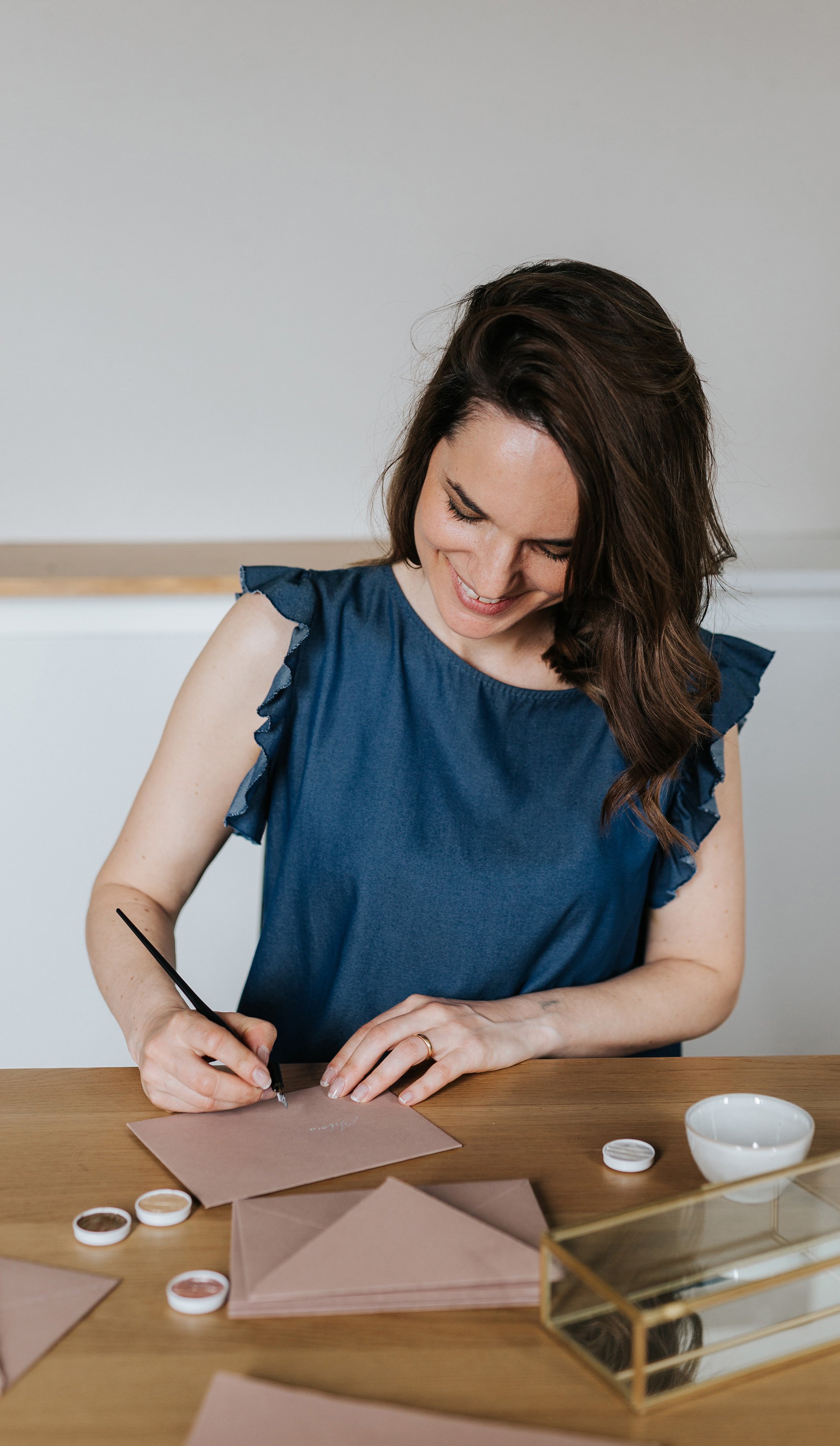 Una donna sorridente seduta a un tavolo crea buste calligrafate a mano.
