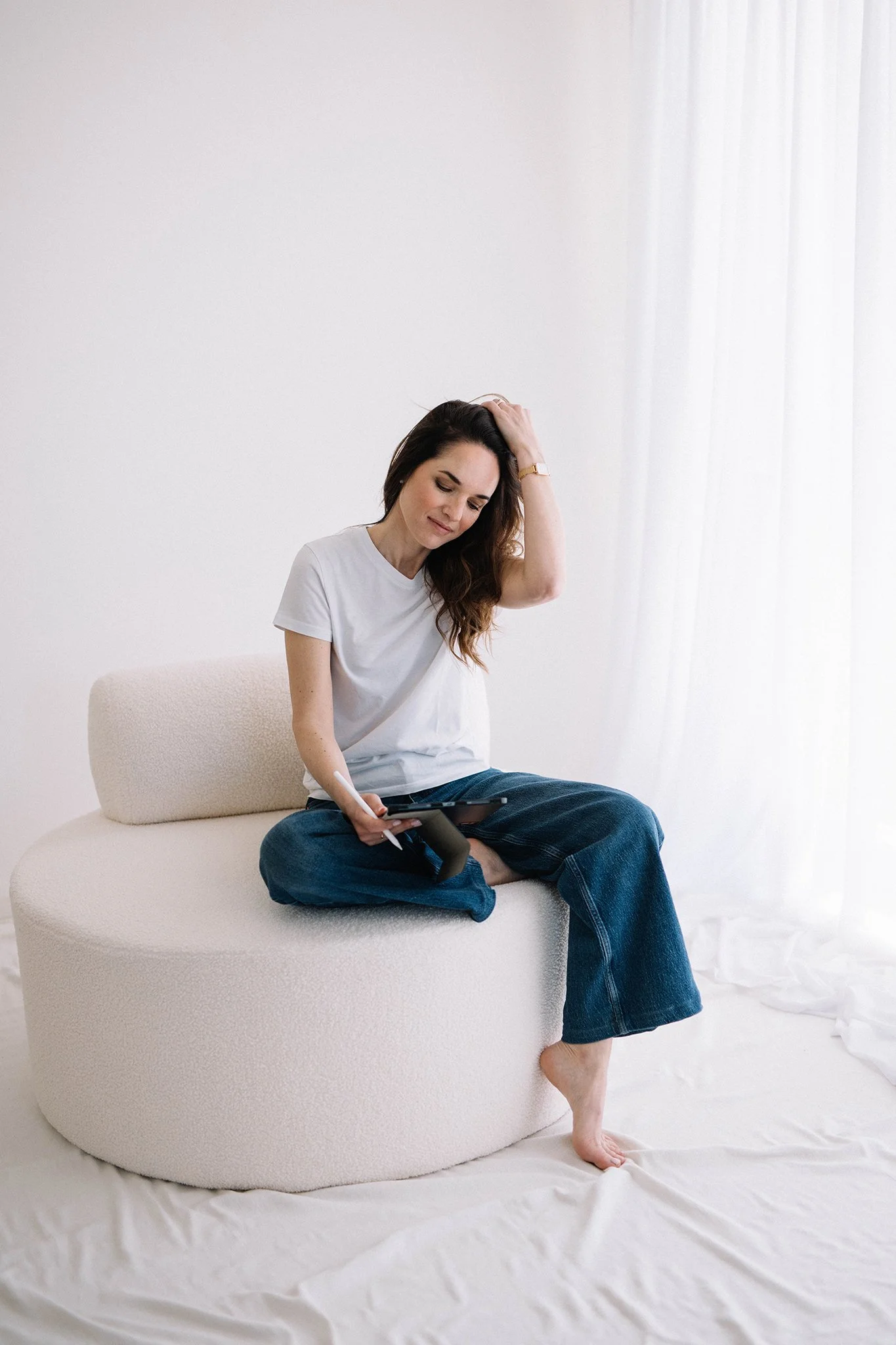 Una donna seduta su un divano, che indossa una maglietta bianca e jeans blu, con una mano sulla testa e l'altra che tiene un tablet, in un ambiente minimalista con pareti bianche, cuscino beige e luci naturali.