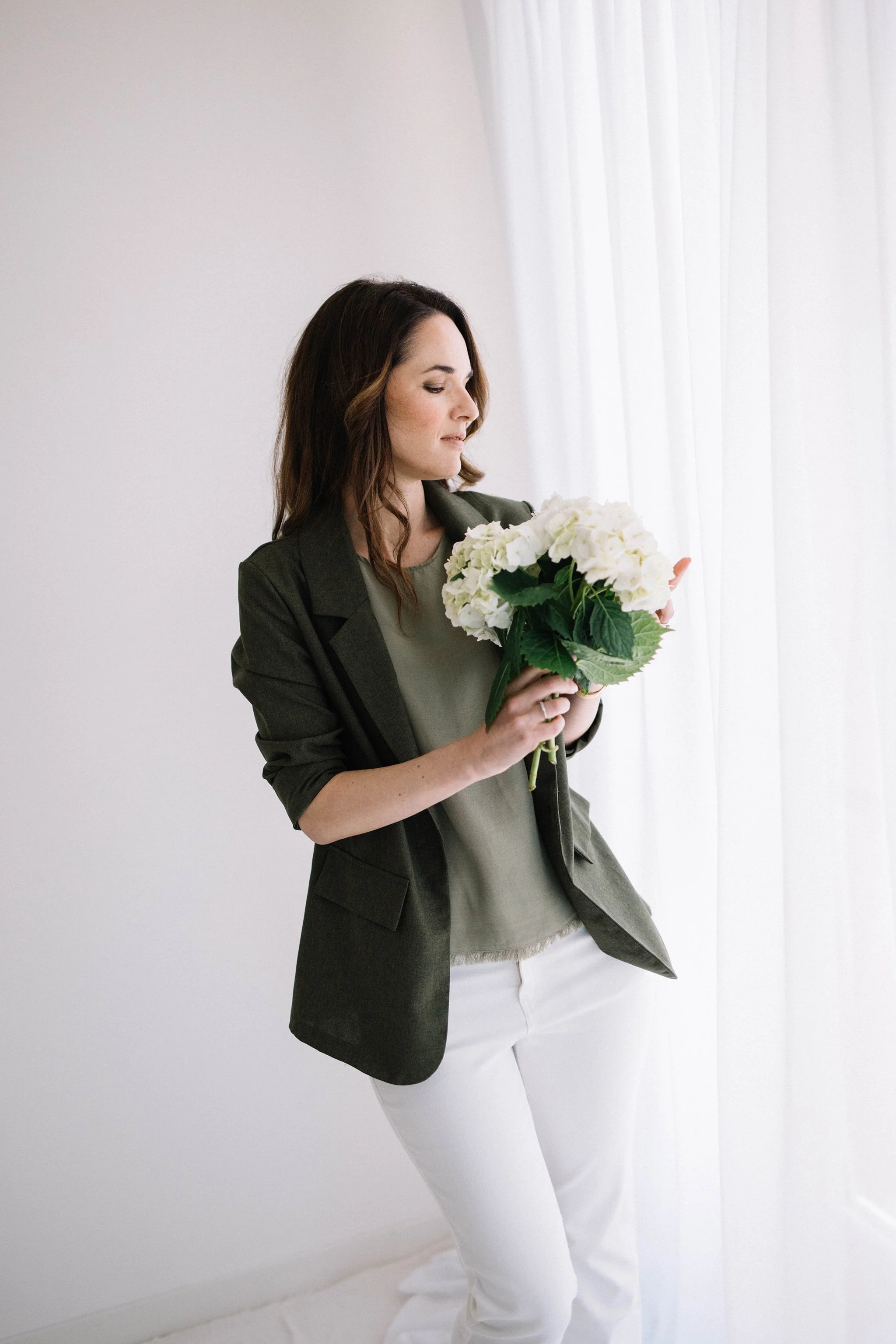donna con capelli castani e vestito verde scuro, che tiene un mazzo di fiori bianchi, in piedi vicino a una finestra con tende bianche, parete bianca sullo sfondo.