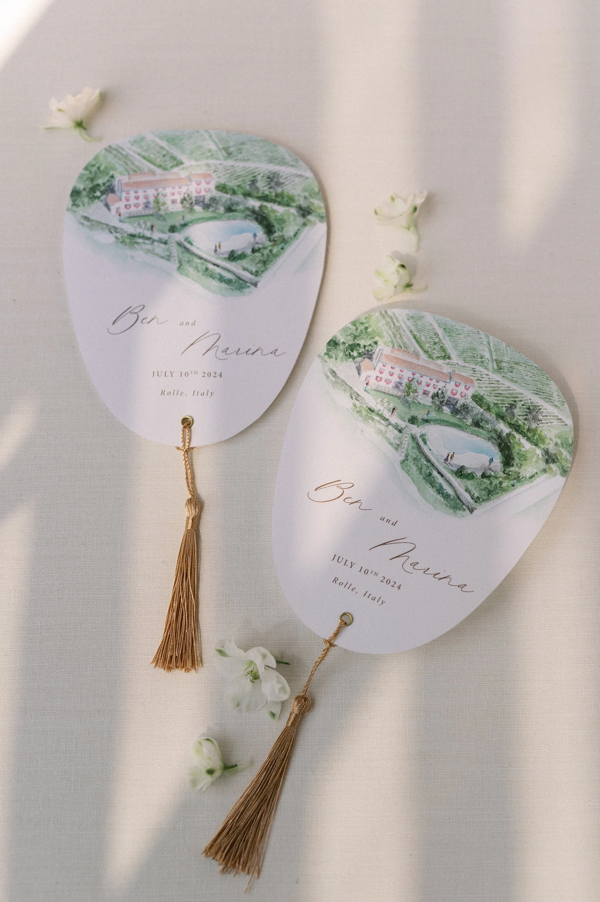 Inviti di nozze con disegno di un paesaggio rurale con edificio, laghetto e vigneti, decorati con fiori e nappe di torsione dorate, con scritte in caratteri eleganti: Ben and Marina, 10 luglio 2024, Rolle, Italia.