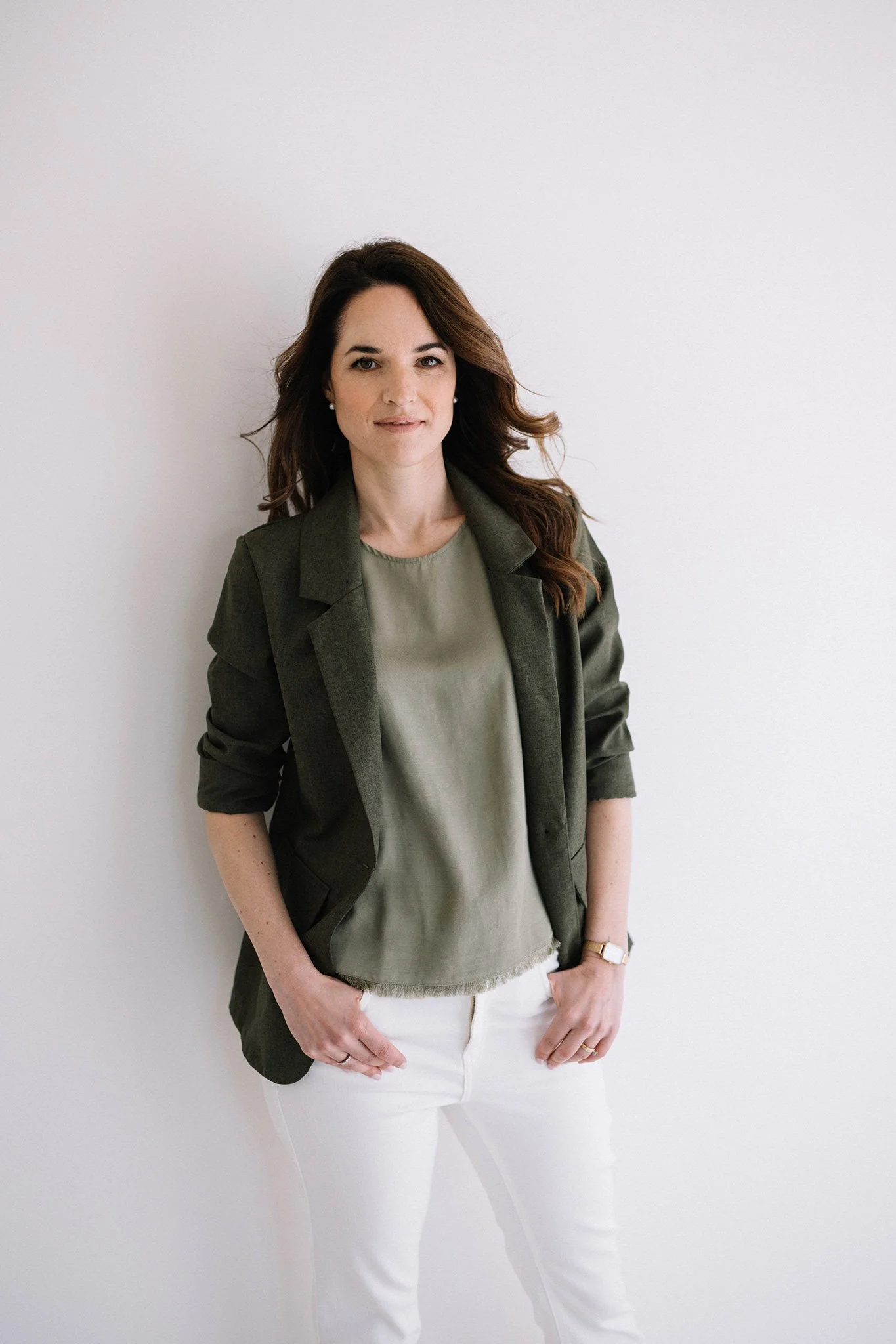 Donna con capelli castani riccioli, indossa una giacca verde scuro, blusa beige e pantaloni bianchi, posa contro una parete bianca.