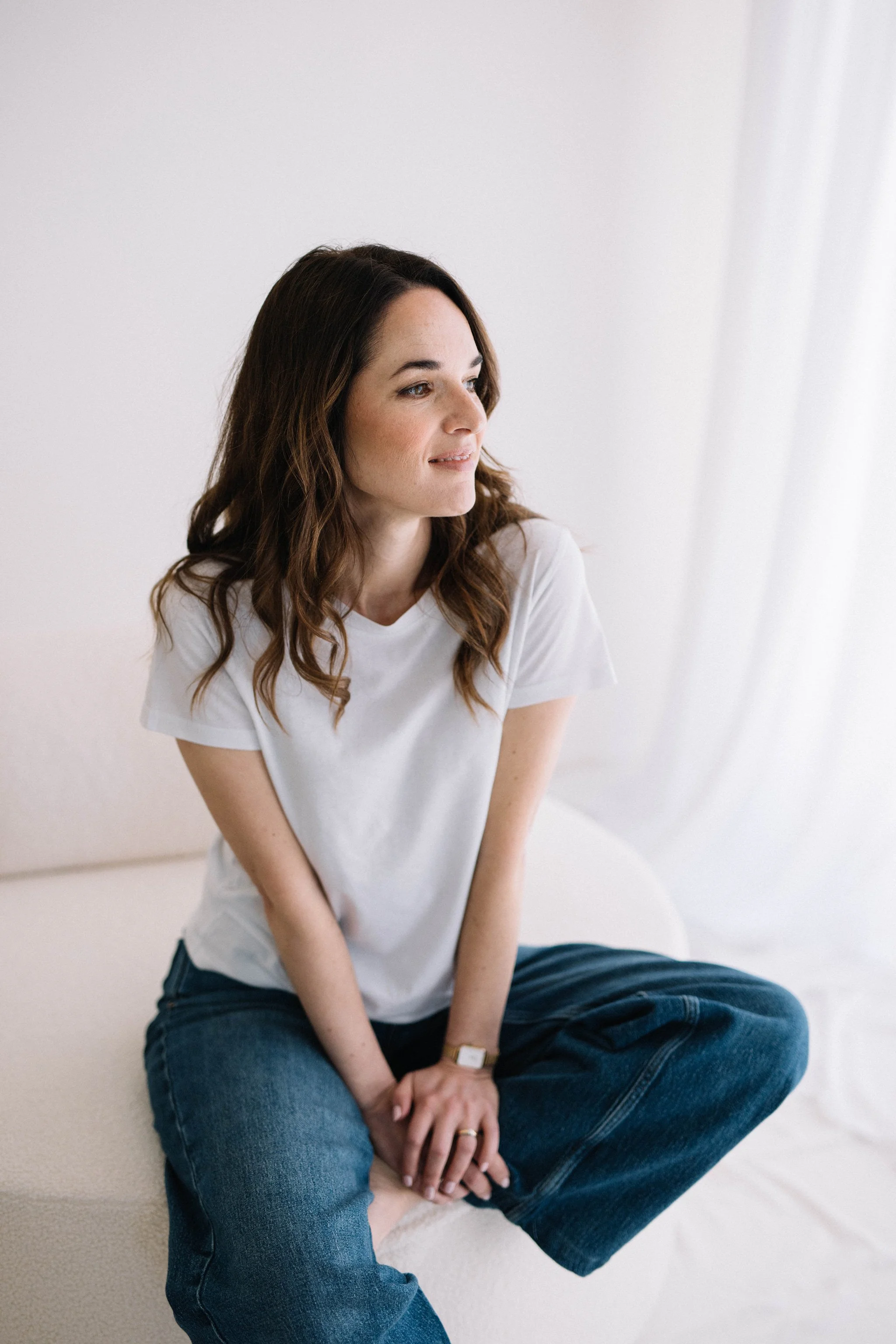 Una donna con capelli marroni ondulati, occhi chiari, vestita con una maglietta bianca e jeans blu, seduta con le gambe incrociate su un tappeto chiaro vicino a un muro bianco.