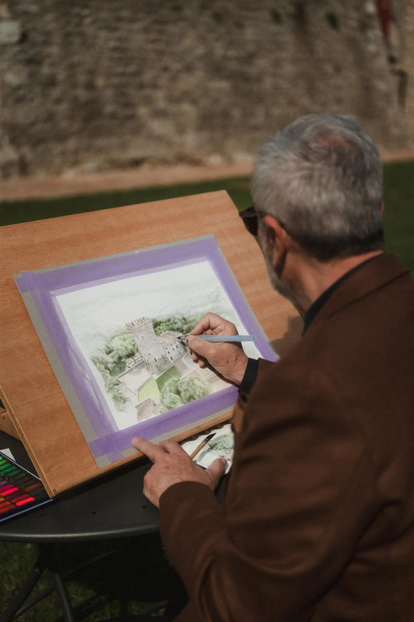 Uomo con capelli grigi e occhiali che disegna o colori un'illustrazione di un castello su carta, seduto di fronte a un tavolo di legno.