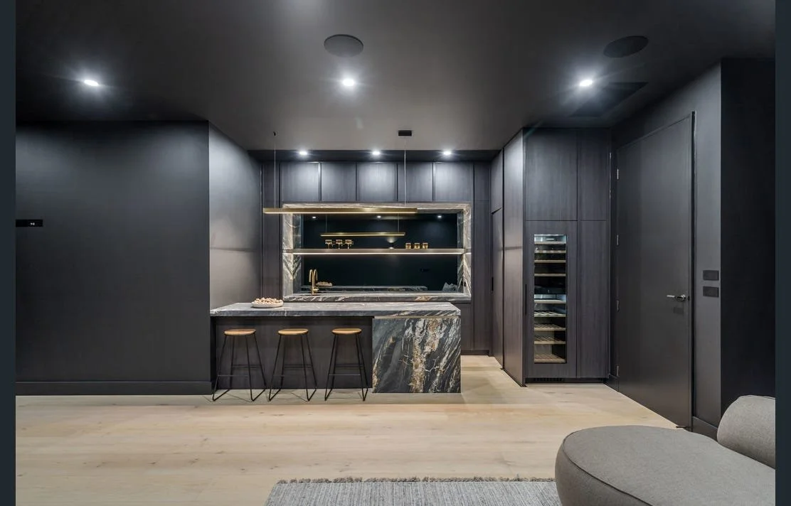 Modern kitchen with dark cabinets, marble countertops, and a built-in wine cooler, illuminated by ceiling lights.