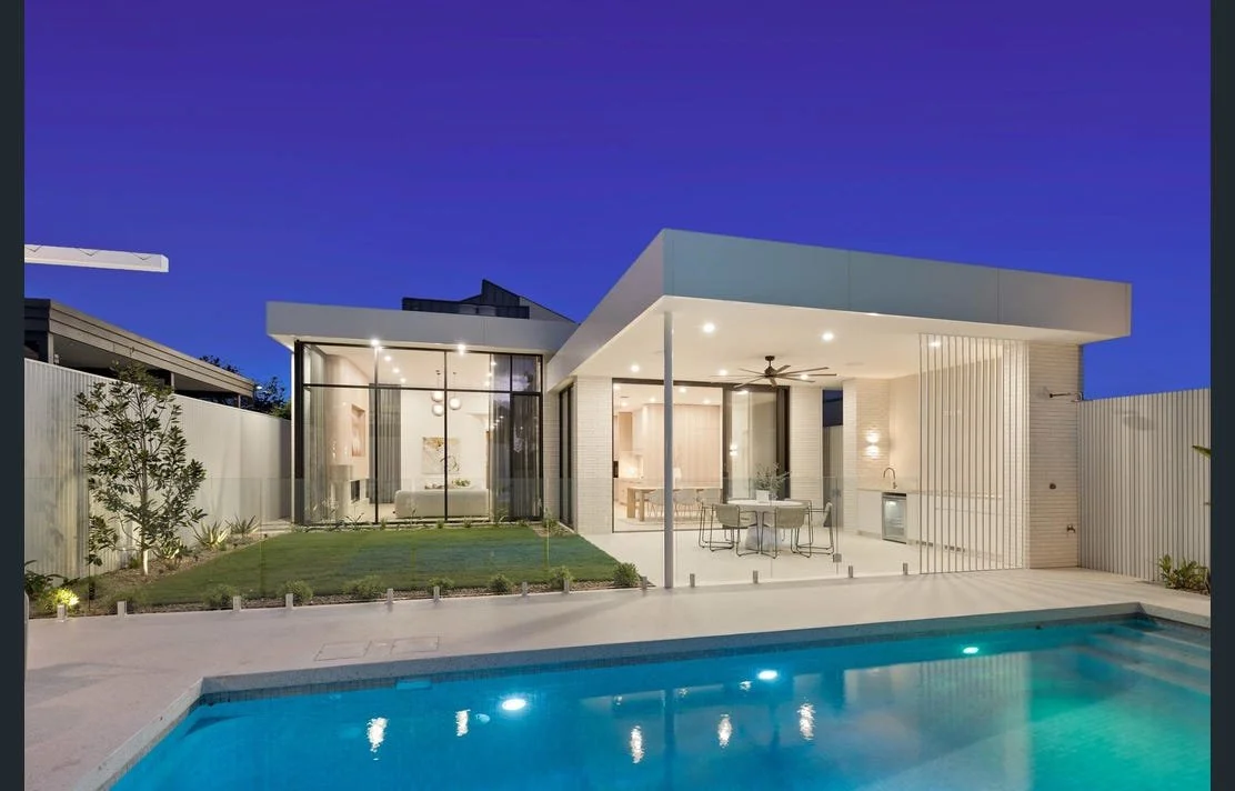 Modern house with glass walls, outdoor patio, and swimming pool at dusk, illuminated interior and exterior lighting.