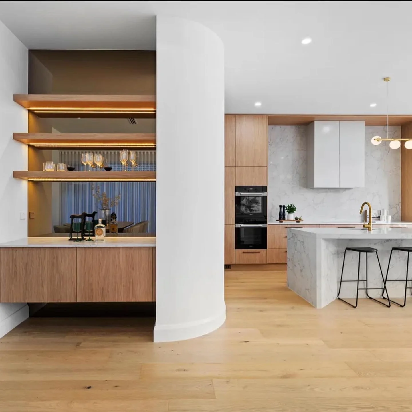 Modern kitchen with wooden cabinets, marble island, gold faucet, and black bar stools, open to dining area with glassware and decorative items.