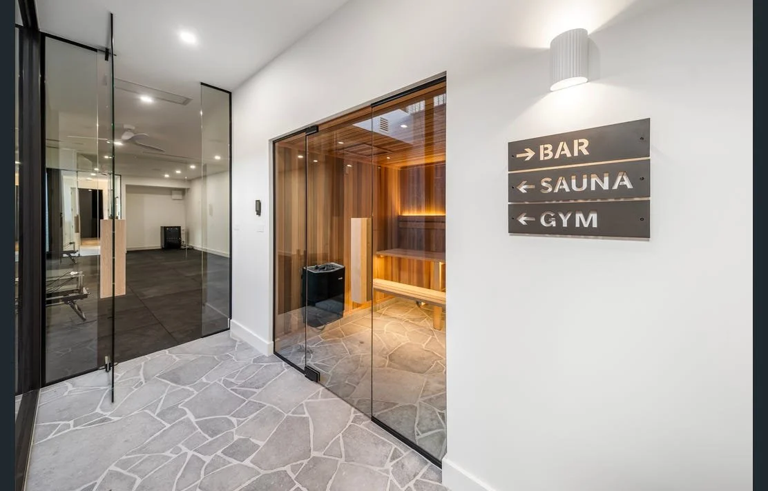 Indoor wellness area with a sauna enclosed by glass walls, adjacent to a bar and gym area. The space features modern decor with stone flooring and white walls, and a sign pointing to the bar, sauna, and gym.