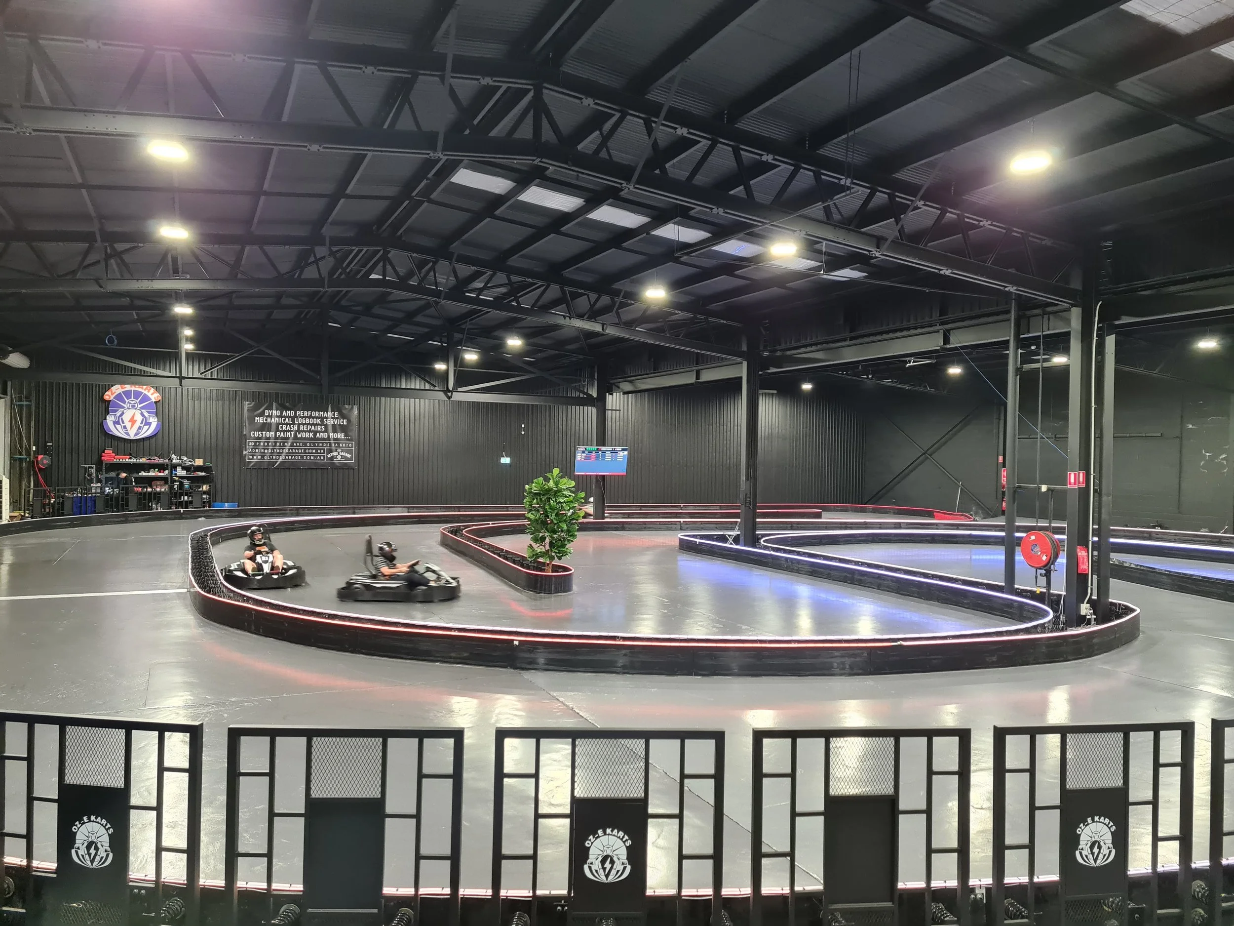 Indoor go-kart racing track with a black and red track layout, multiple racers in go-karts, and a black wall with a logo of Oz-E Karts.