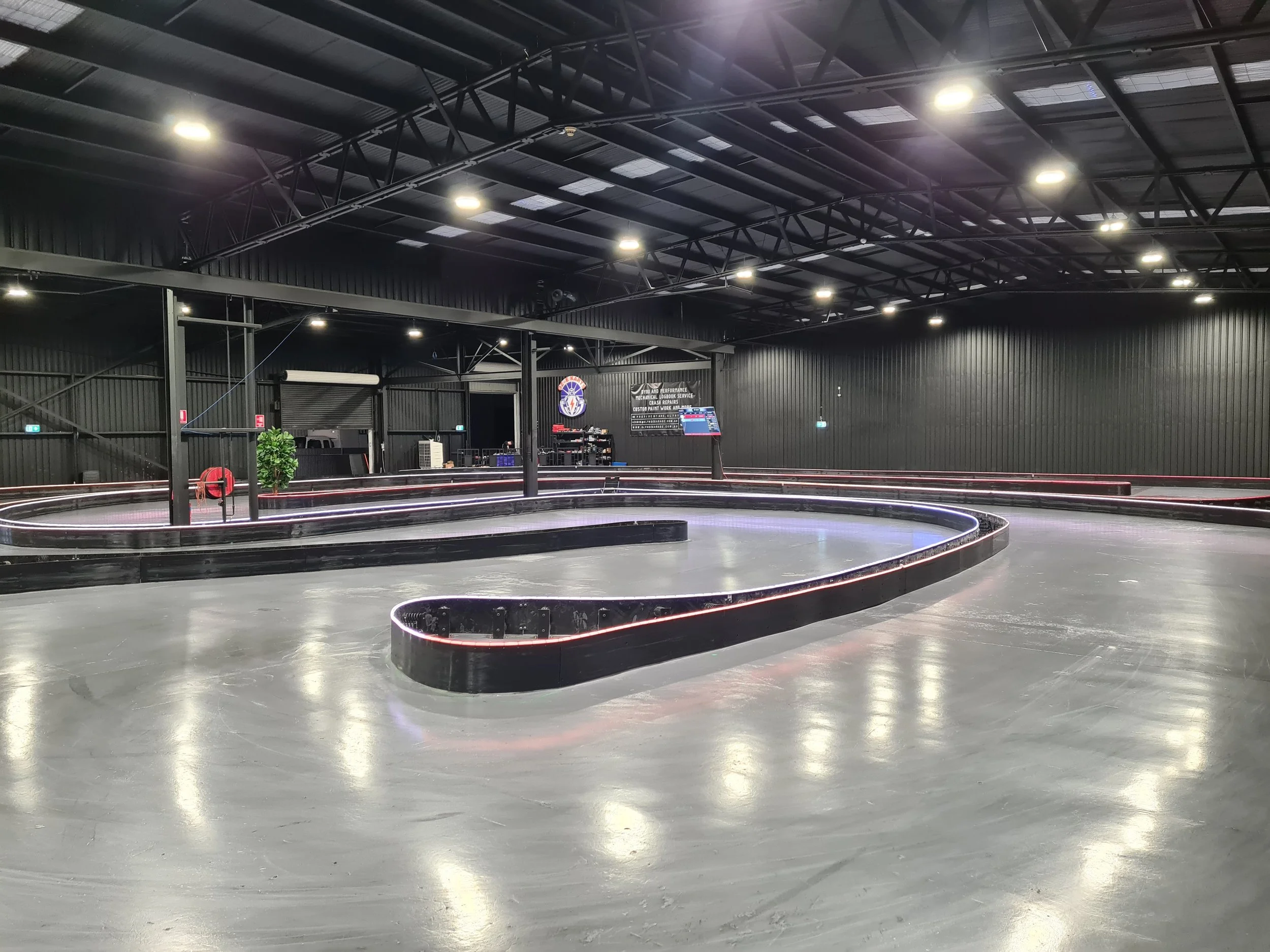 Indoor go-kart racing facility with a zigzag track, black walls, and LED lights on the ceiling, with a sign in the background indicating mechanical, electrical, and laser services advertisement.