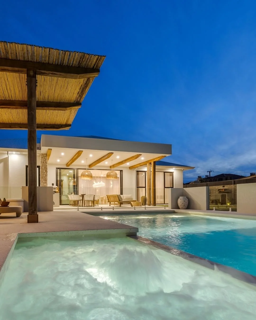 Modern house with outdoor pool and hot tub at dusk, illuminated patio with seating area, glass walls, wooden accents, and bamboo shade structure.