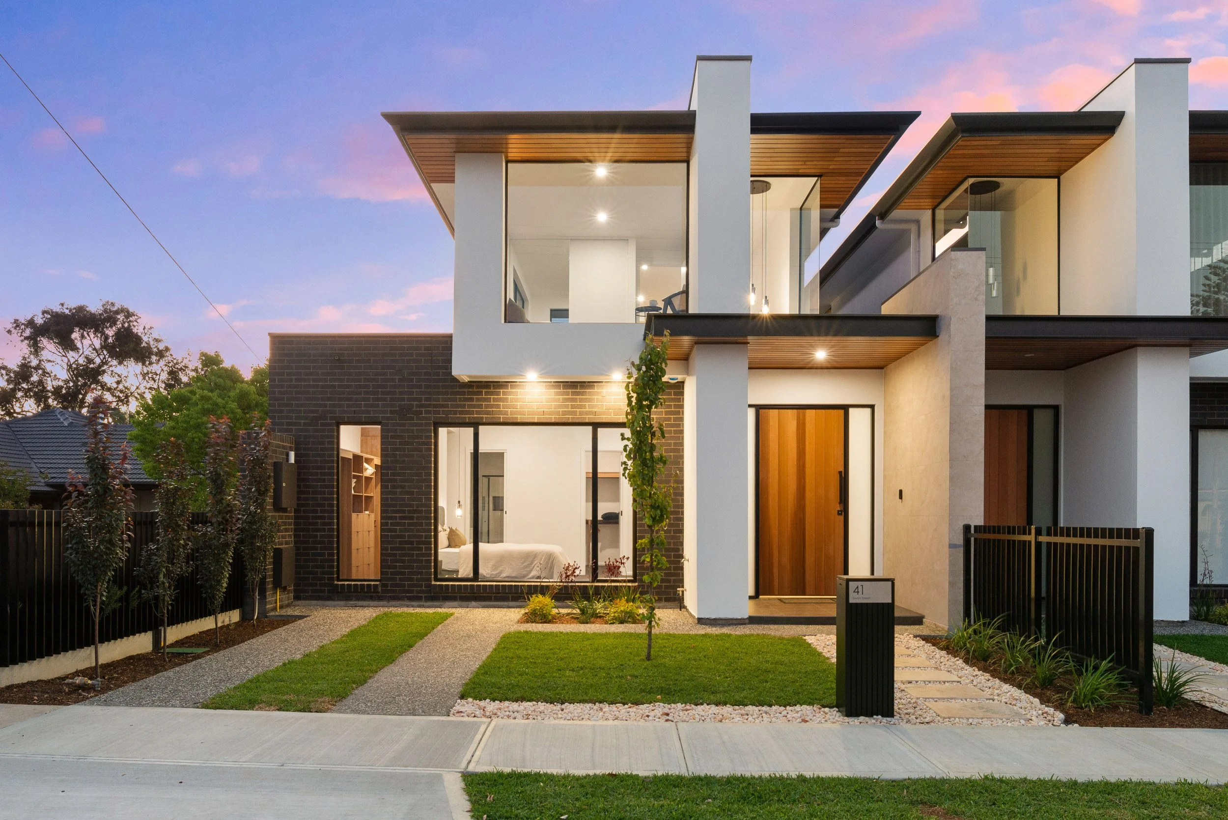 Modern two-story house with large windows, wooden accents, and a landscaped front yard at dusk.