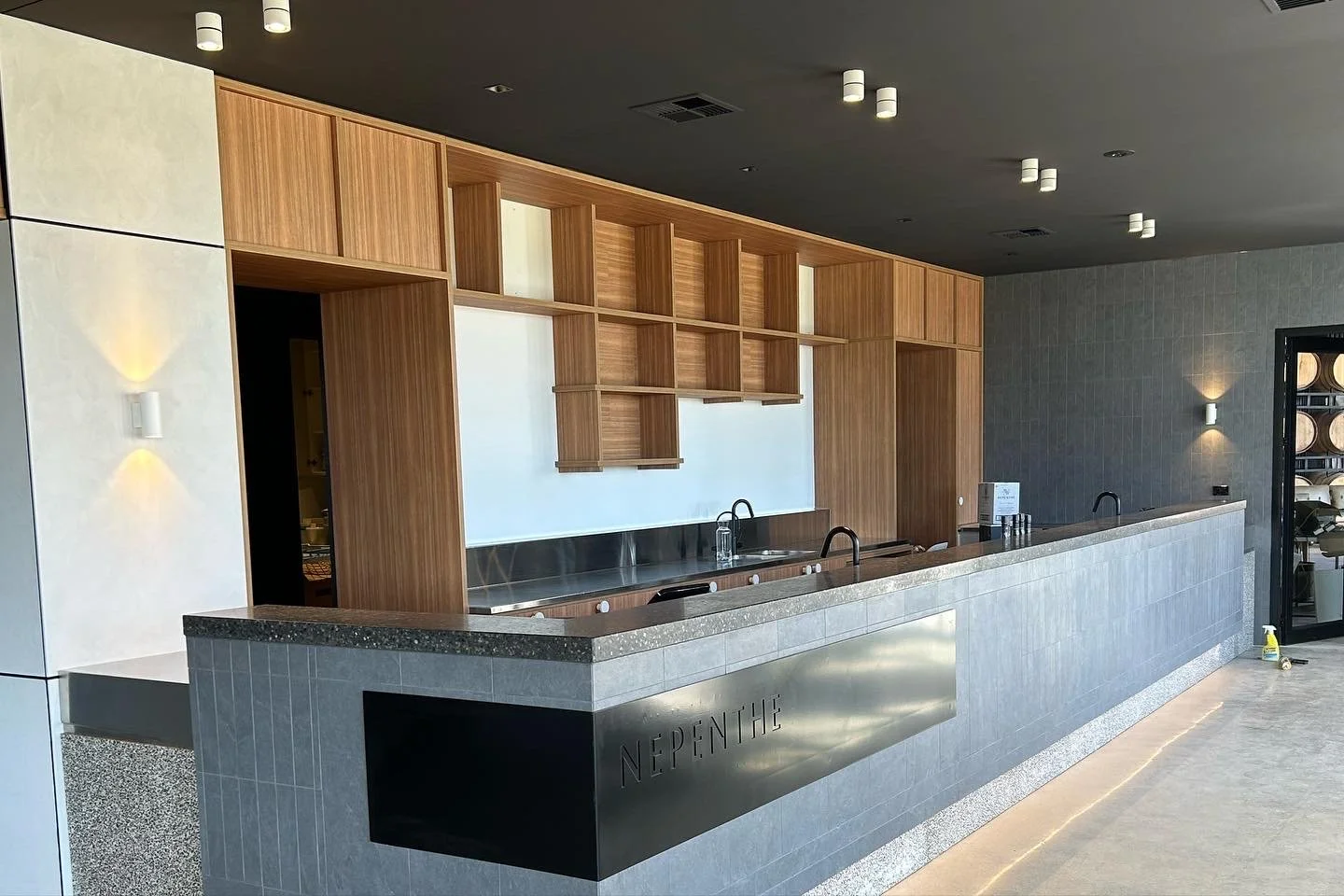Modern coffee bar with wooden shelving and a granite countertop, labeled 'NINE THE' in minimalist style, with a dark ceiling, light fixtures, and subtle lighting on the wall.