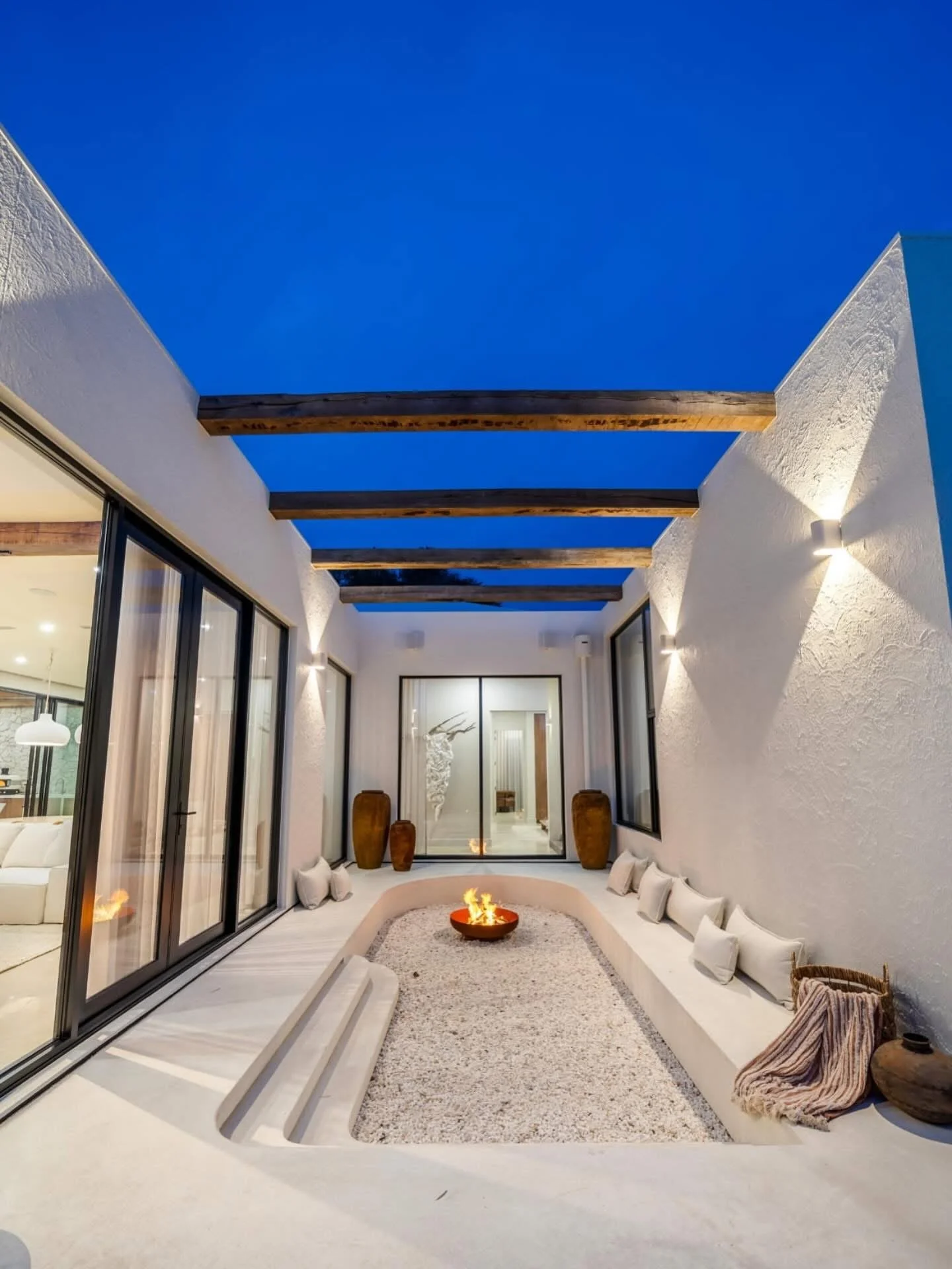 Contemporary outdoor patio with built-in seating, fire pit, potted decor, and large glass sliding doors, under a partially open roof with exposed wooden beams, illuminated by wall sconces