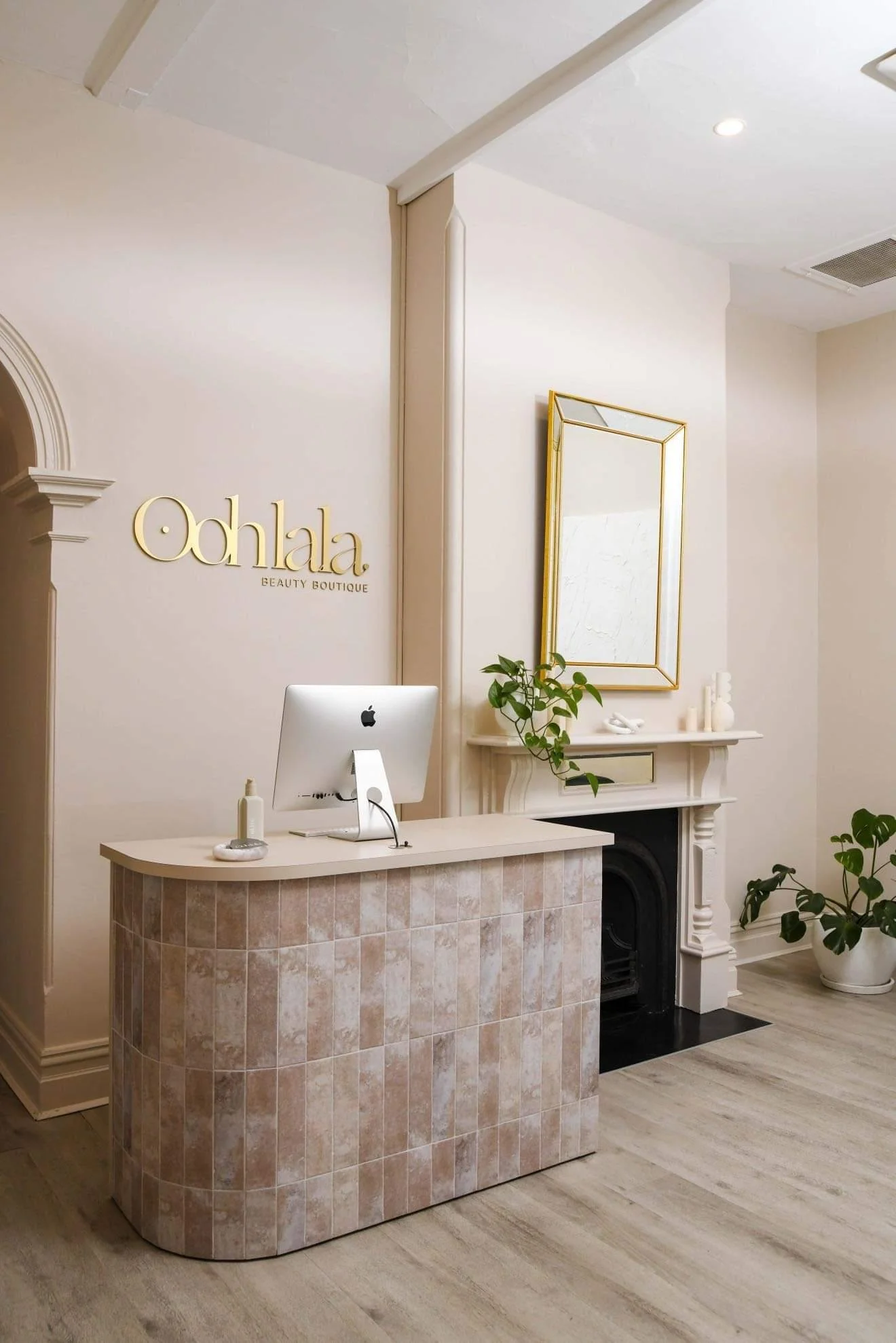 Reception area of a beauty boutique with a white counter, computer, and a mural on the wall reading 'Ohlala Beauty Boutique,' with a decorative mirror, fireplace, and potted plants in a minimalist, elegant style.