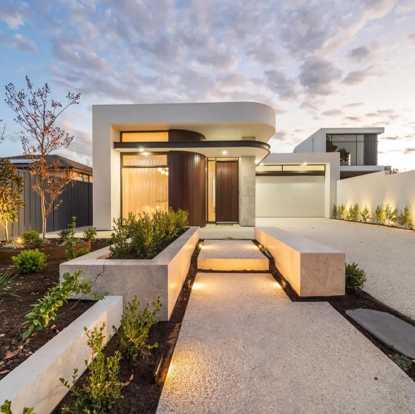 Modern house with glass and wooden door, landscaped front yard with plants and a pathway, evening lighting with a partly cloudy sky.