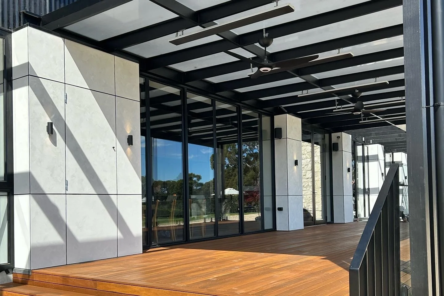 Modern outdoor patio with wooden flooring, black metal beams, ceiling fans, and large glass windows.