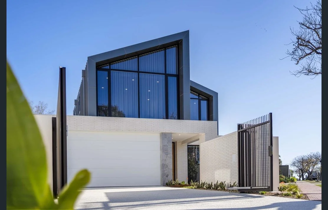 Modern two-story house with large glass windows, a white brick facade, and a black metal gate, with a clear blue sky and trees in the background.