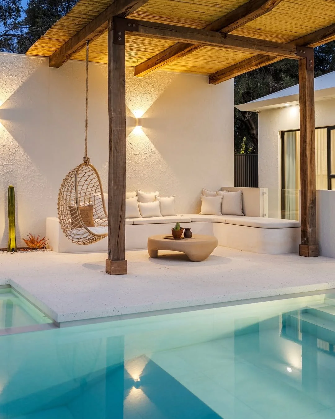 A modern outdoor patio area with a swimming pool, featuring a built-in lounge with beige cushions, a round coffee table with a small plant and pottery, a hanging wicker chair, and a wooden pergola with warm lighting against white stucco walls.