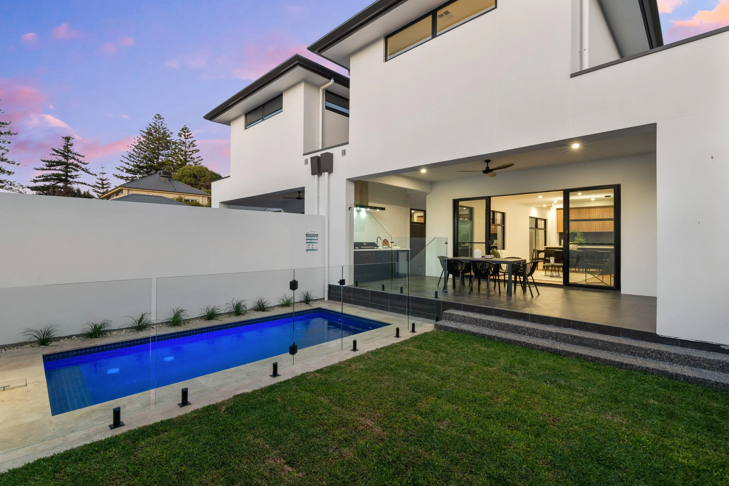 Modern white multi-story house with large windows, an outdoor patio, a swimming pool with blue lighting, and glass fencing, during dusk with a colorful sky and trees in the background.