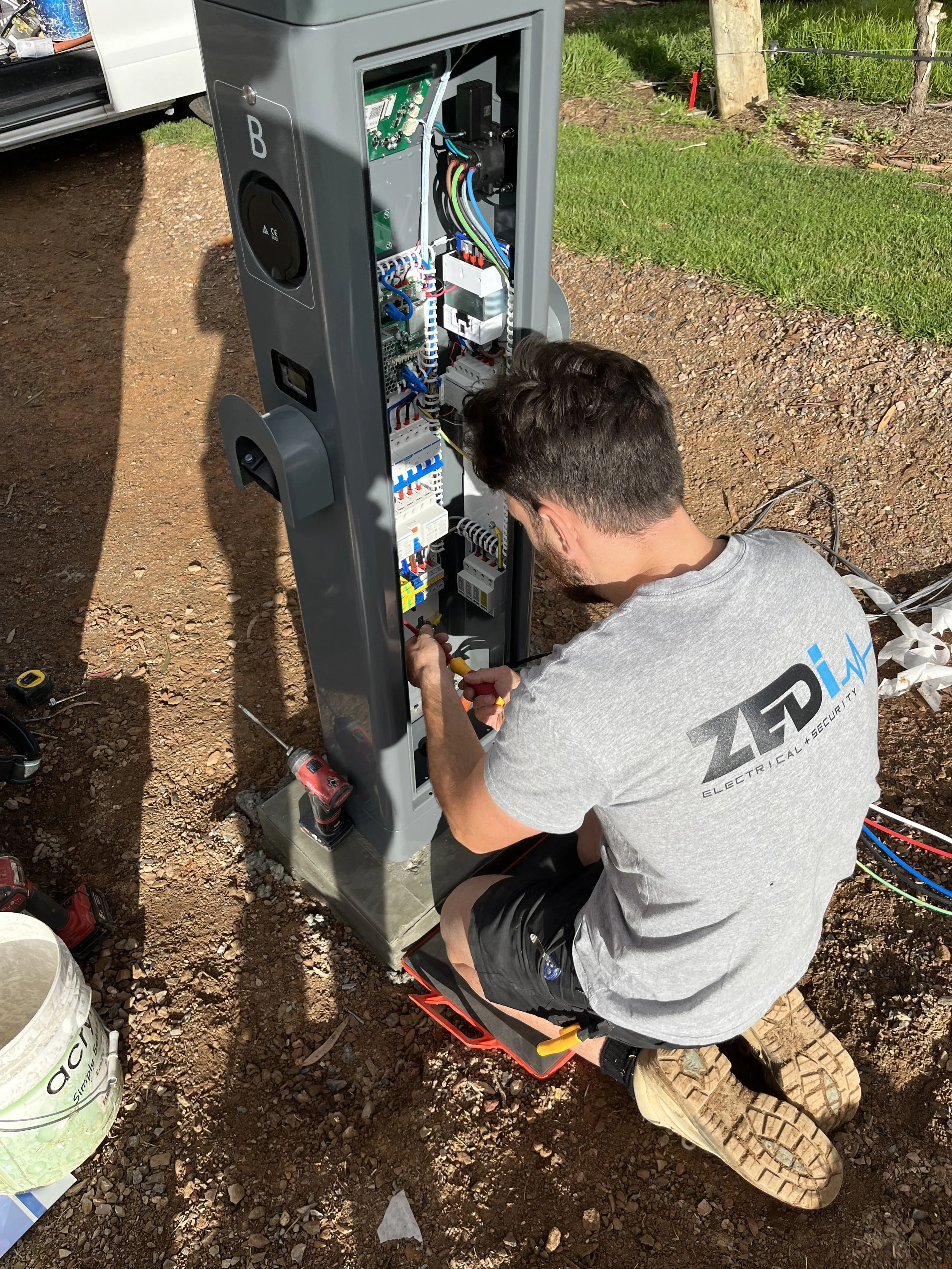 A technician working on an outdoor electrical control panel, kneeling on the ground with tools, surrounded by various equipment and wiring, with grass and dirt ground nearby.