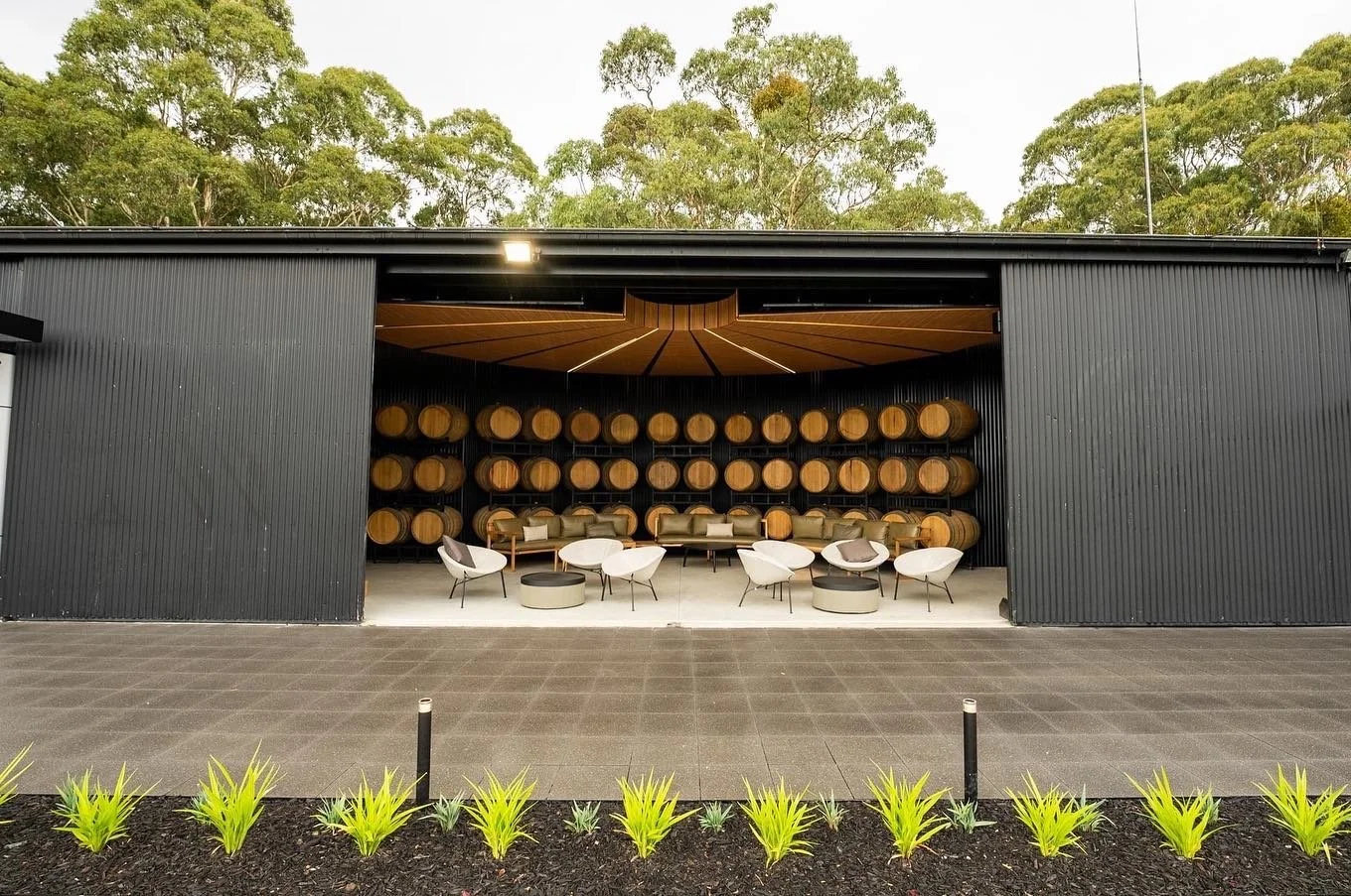 Outdoor winery seating area with black building, wine barrels on shelves, white chairs, and green plants in front.