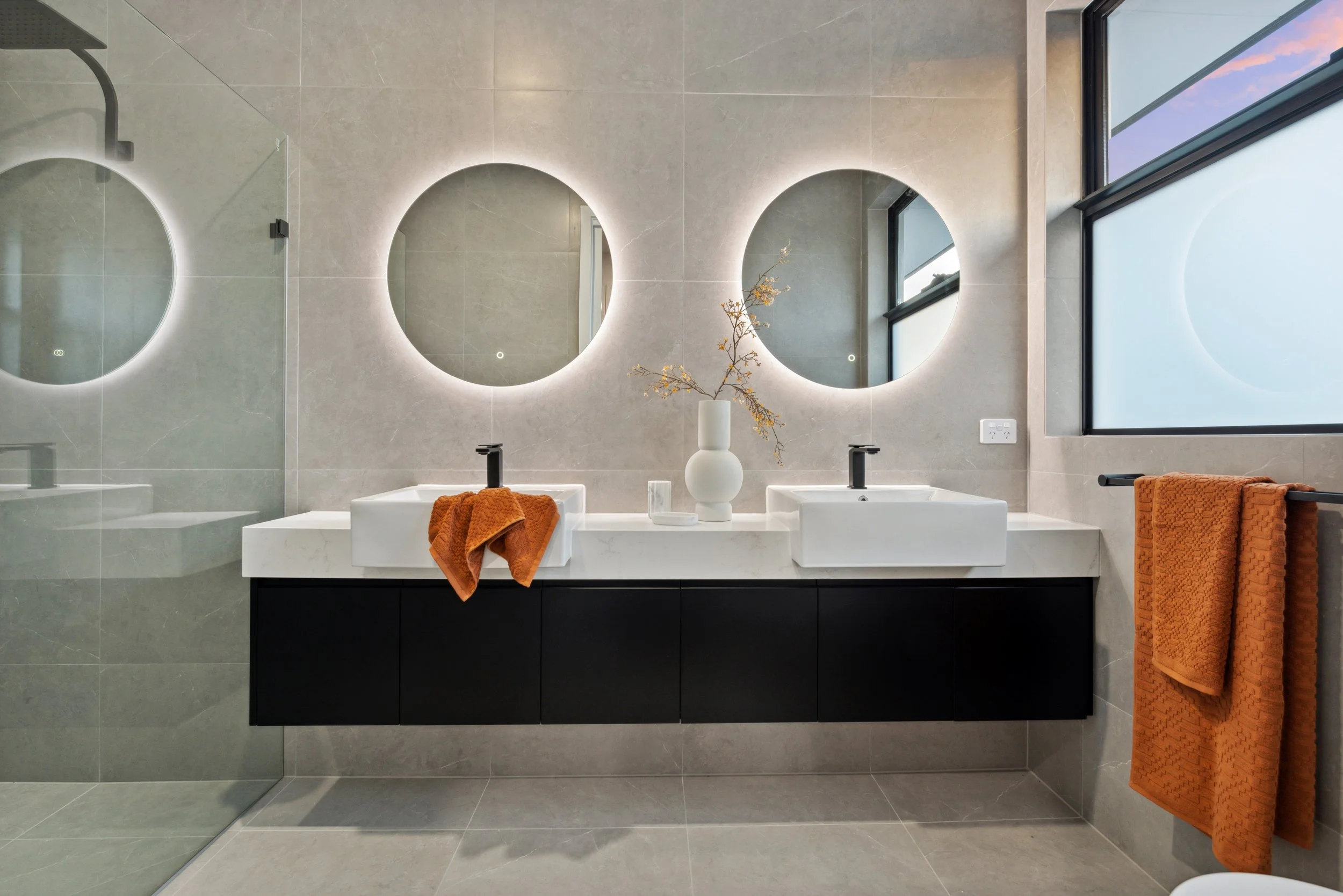 Modern bathroom with double sinks, round backlit mirrors, large window, orange towels, and a minimalist decor with a vase and decorative branches.