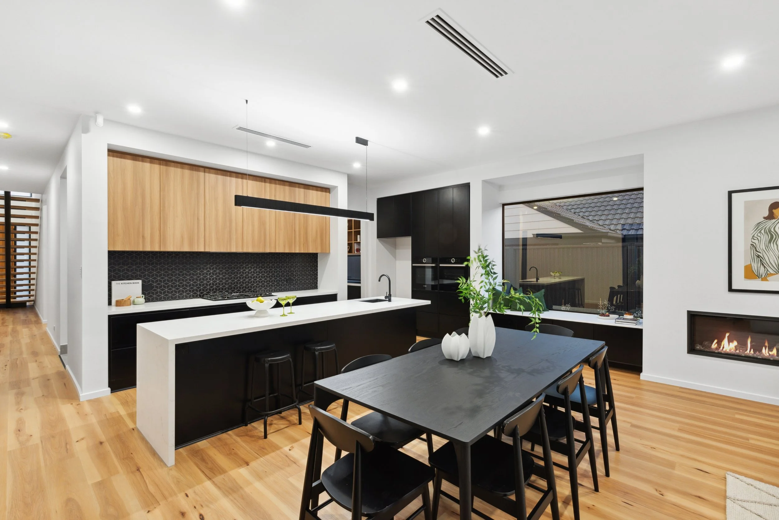 Modern open-concept kitchen and dining area with black and wood cabinets, a white kitchen island, and a dining table with black chairs. A large window reveals a tiled roof outside. The ceiling has recessed lighting and black linear light fixtures. The space has wooden flooring and contemporary decor.