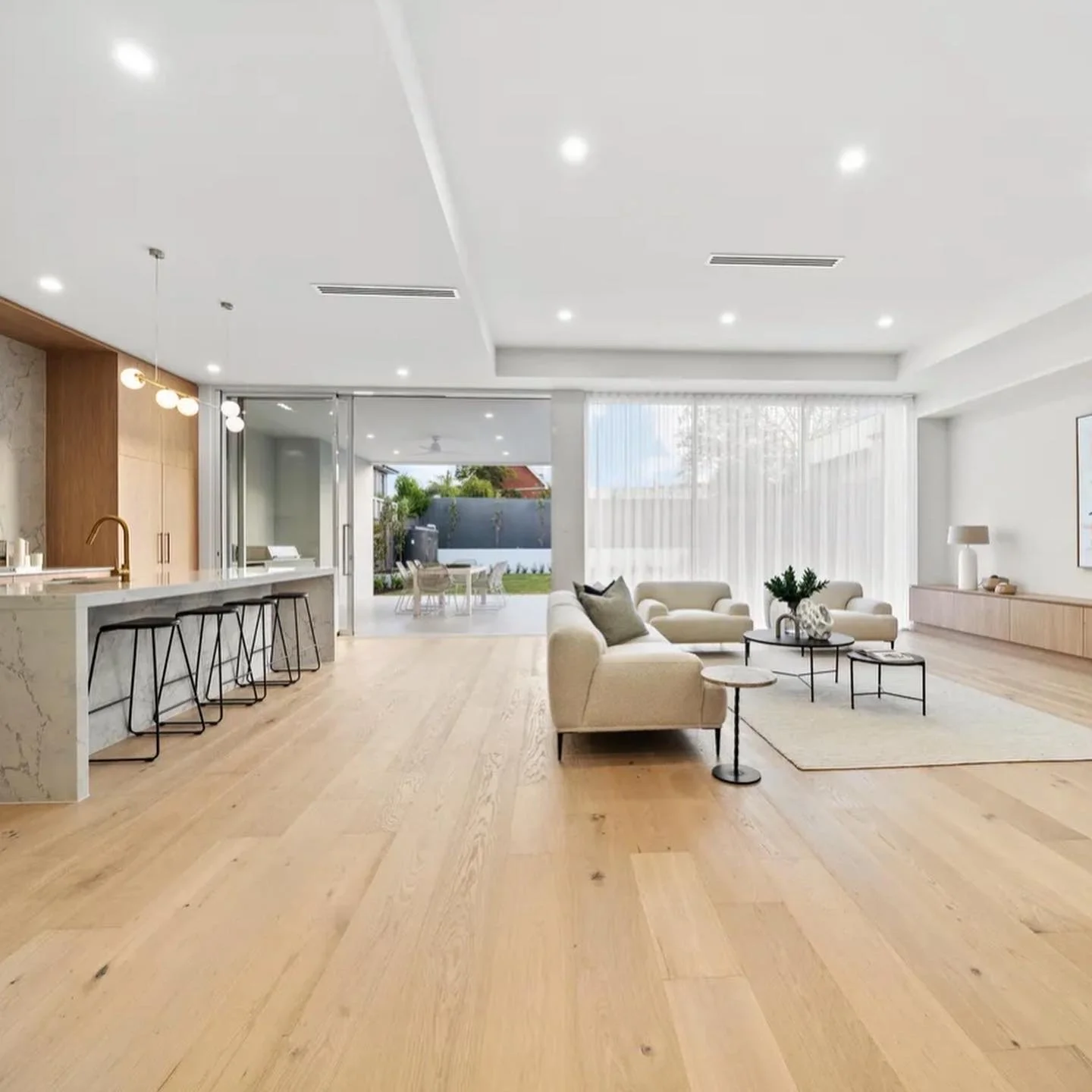 Modern open-concept living room with white sofas, black coffee tables, and a view of the outdoors through large windows, featuring light wood flooring and minimalistic decor.