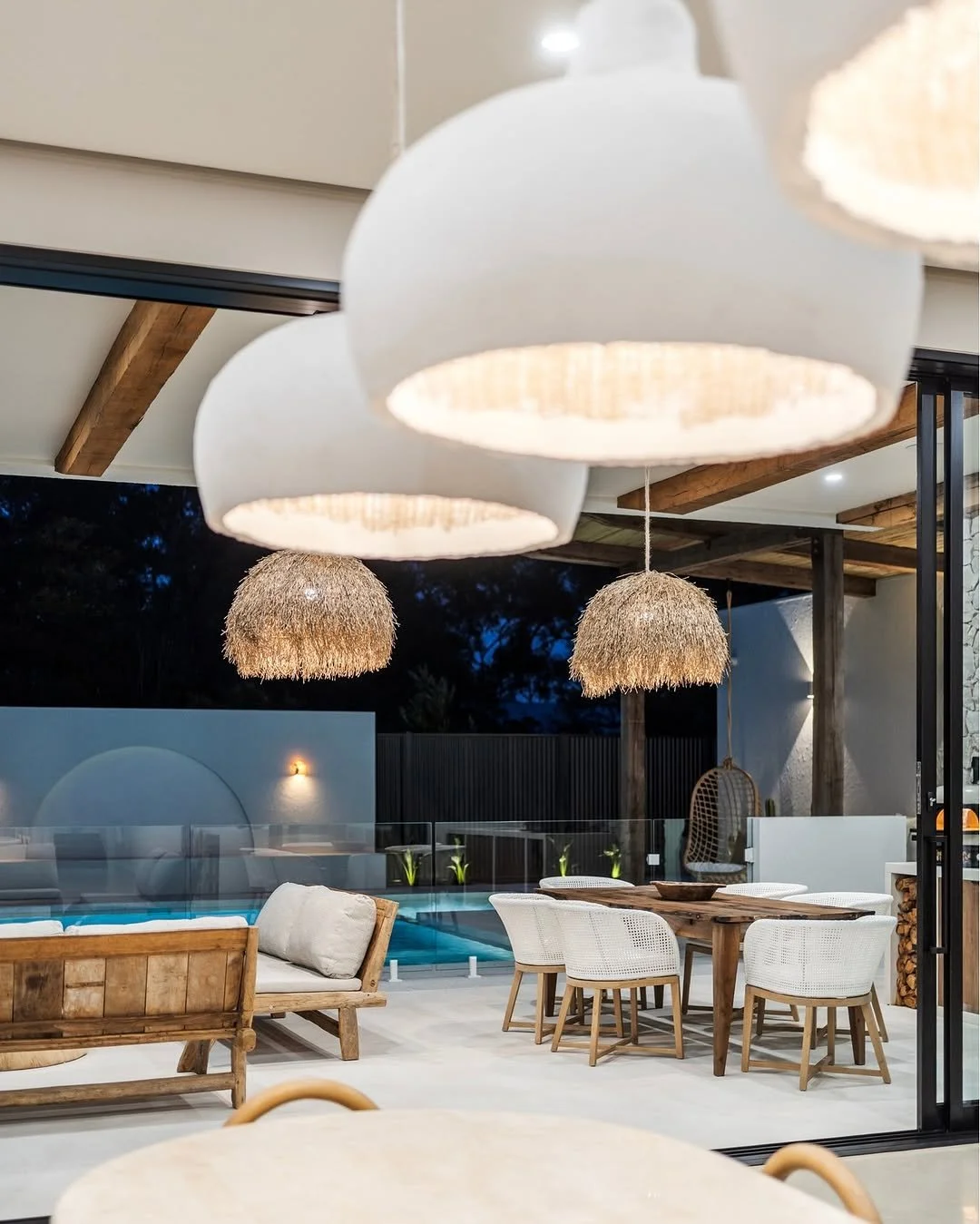 An outdoor patio area at night with hanging pendant lights, a wooden outdoor dining table with six white chairs, a wooden bench with cushions, and a swimming pool in the background surrounded by a glass fence.