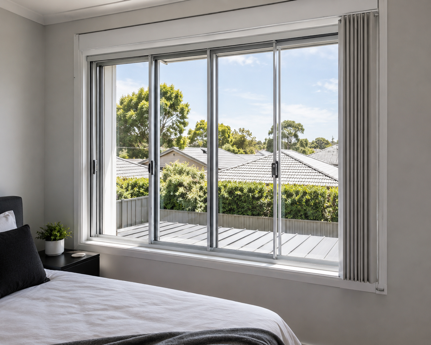 A bedroom with a large sliding glass window overlooking a backyard with trees and neighboring rooftops. The bed has white linens and black pillows, with a small black nightstand and a potted plant next to it.