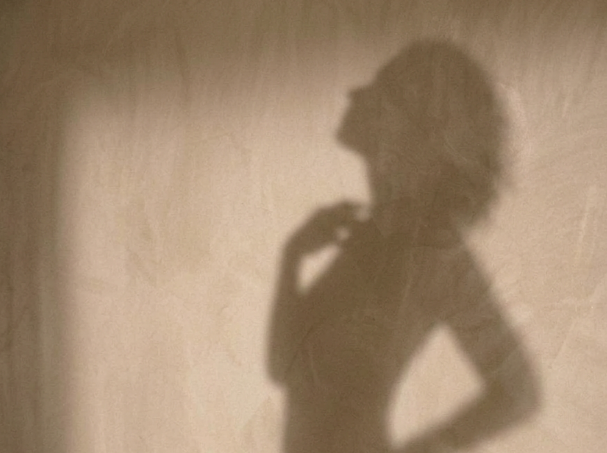 Shadow of a person with curly hair posing with one hand on their hip and the other hand near their chin, cast on a plain wall.