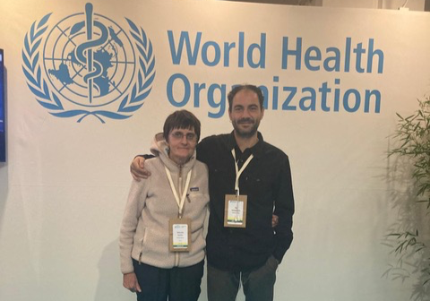 Due persone, un uomo e una donna, sorridenti, si abbracciano davanti a un cartellone con il logo dell'Organizzazione mondiale della sanità, durante un evento.