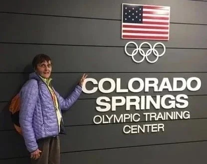 Una persona davanti a un muro con il segno 'Colorado Springs Olympic Training Center', una bandiera degli Stati Uniti e i cerchi olimpici.